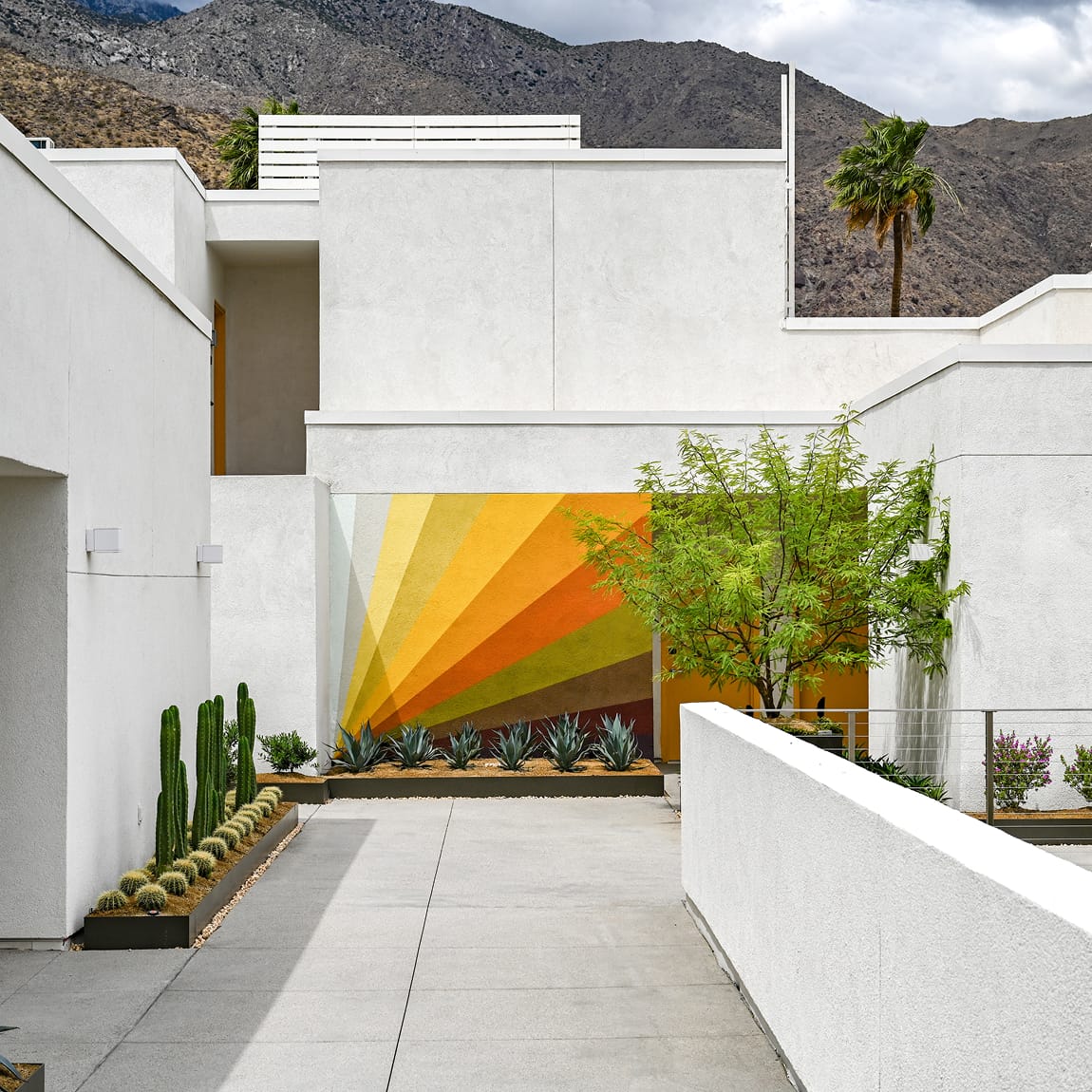 Outdoor courtyard corridor at Thompson Palm Springs with white stucco architecture, a sunburst mural in warm yellow and orange tones, desert plantings of agave and columnar cacti, and the San Jacinto Mountains visible in the background.