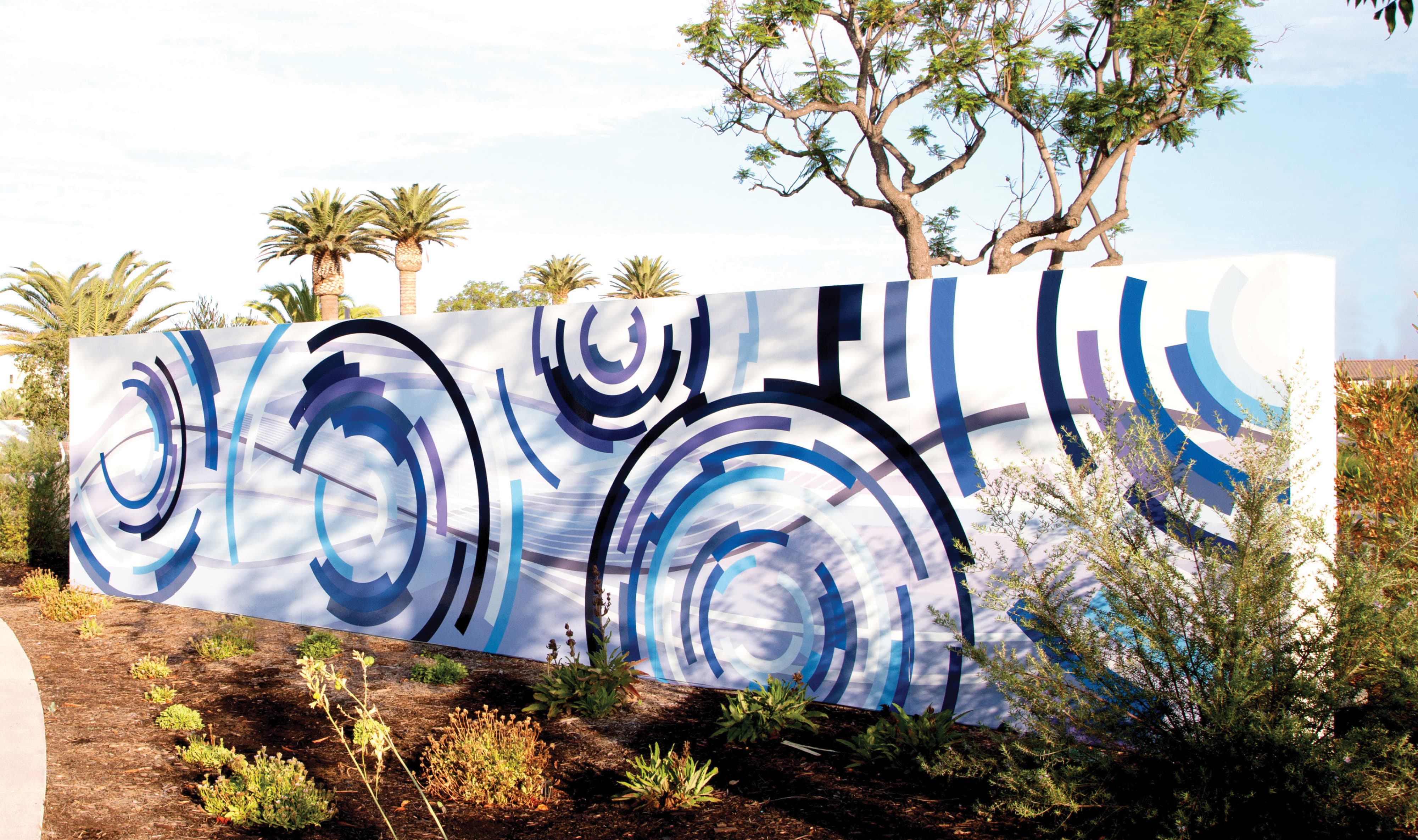 Freestanding art wall with a mural of overlapping blue, navy, and purple concentric circles, set among palm trees and desert landscaping along a garden path.