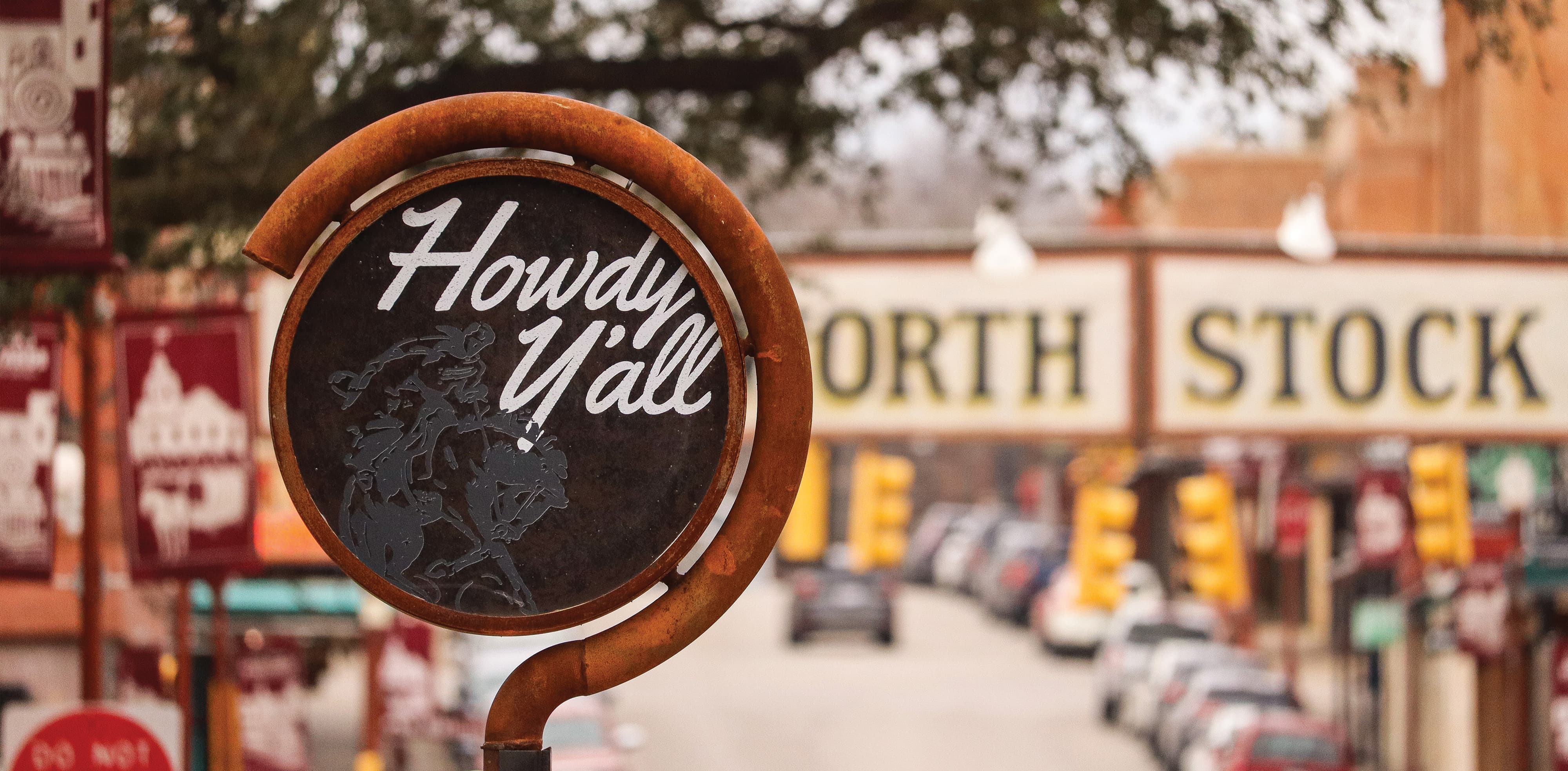 A circular sign with the text “Howdy Y’all” in a casual script font and a faded image of a cowboy on horseback. The sign face is made of a dark, weathered metal. It is suspended from a weathered, rust-orange industrial pipe that has been bent into a semicircle around the sign face, then extends straight down out of frame. Blurred in the distance behind the sign, there’s a vibrant streetscape.