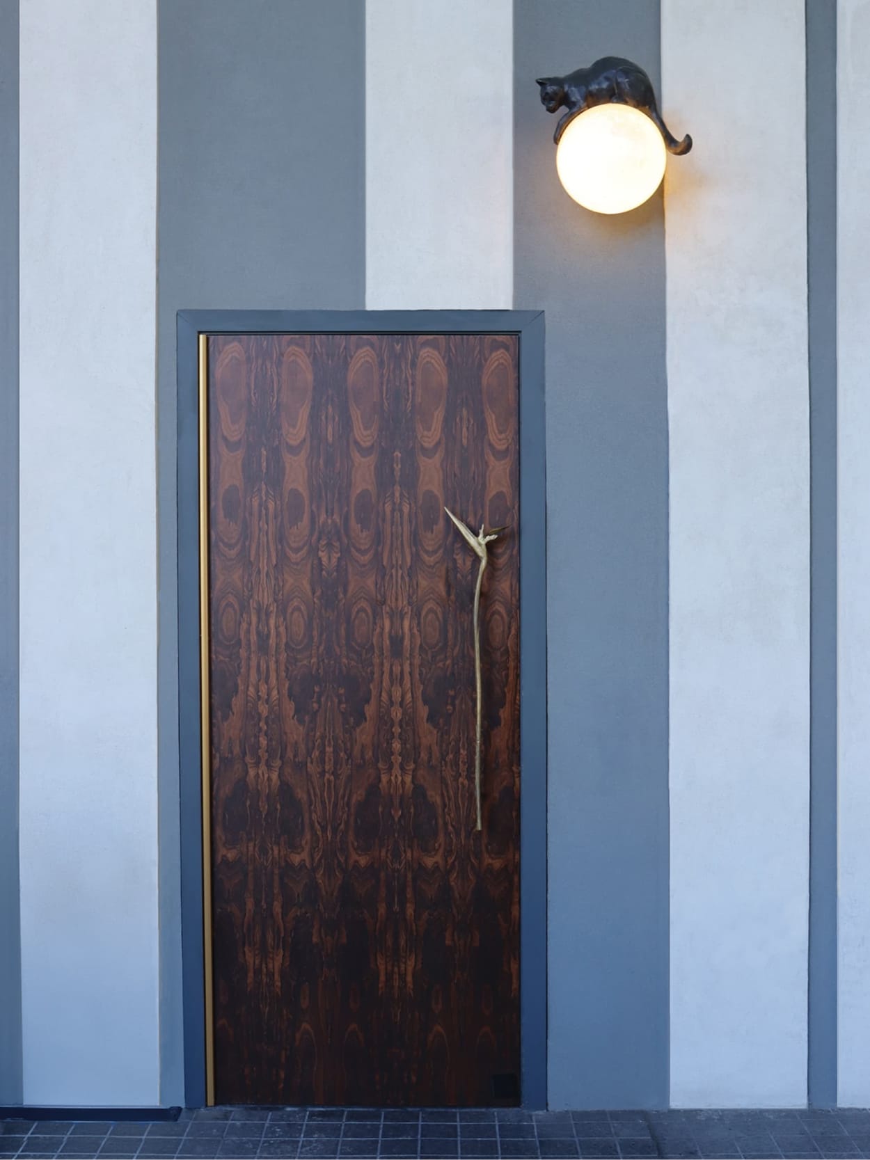 Wood-grain door with a bronze branch handle beneath a globe sconce topped by a cat sculpture at The Swexan Hotel - environmental graphic design by RSM Design