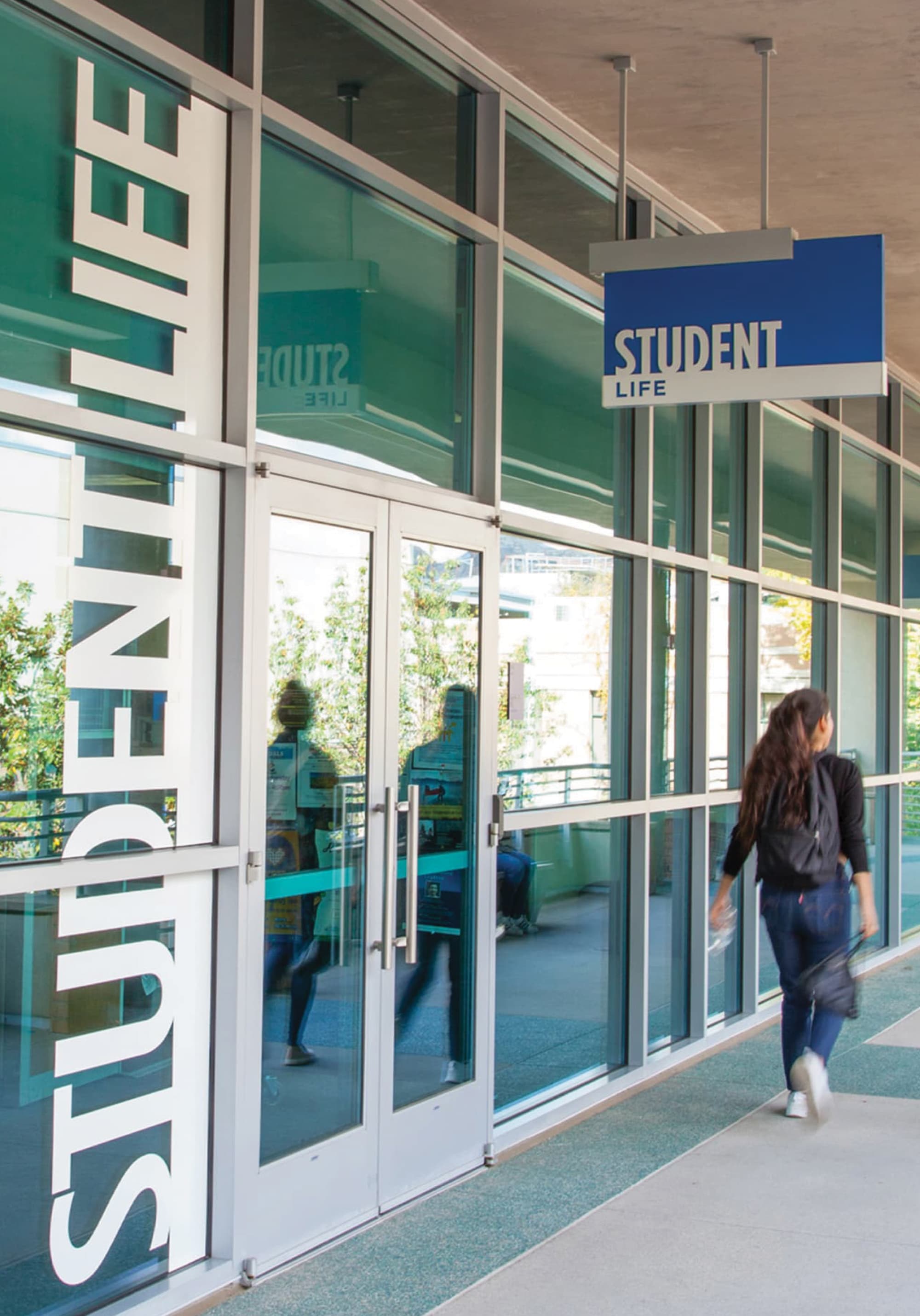 Glass entrance to 'Student Life' with large vertical text on the window and a blue hanging sign, as a student walks along the covered walkway outside.