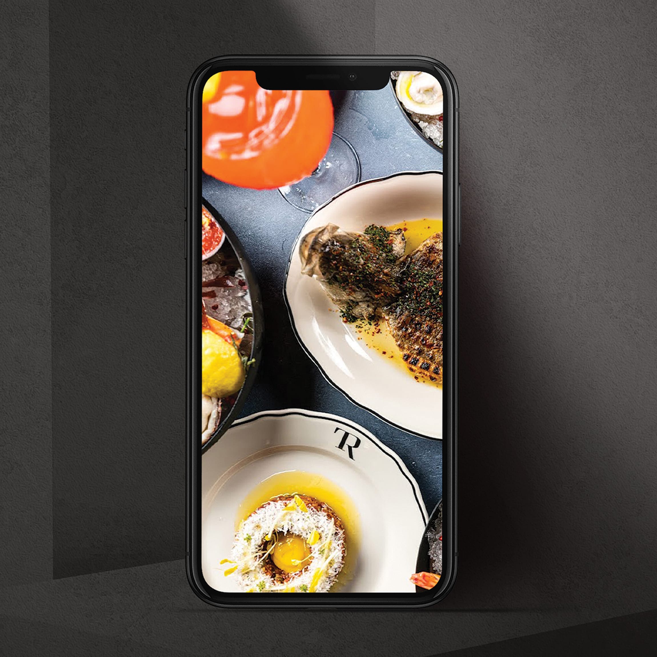 Smartphone displaying overhead photo of plated dishes on monogrammed dinnerware with cocktail and oysters, Trick Rider, Omni PGA Frisco Resort, brand identity by RSM Design.