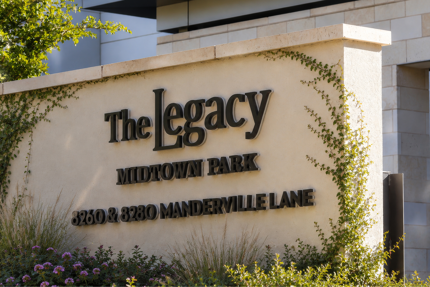 Stone monument sign reading 'The Legacy Midtown Park, 8260 & 8280 Manderville Lane,' covered in climbing vines with flowering shrubs and grasses at its base.