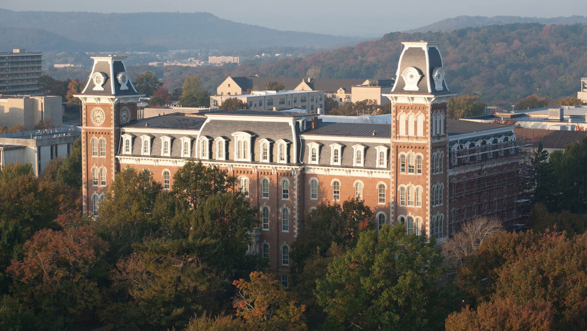 Aerial view of Old Main building at the University of Arkansas in Fayetteville featuring iconic twin clock towers, red brick Second Empire architecture, and fall foliage with Ozark Mountains in background