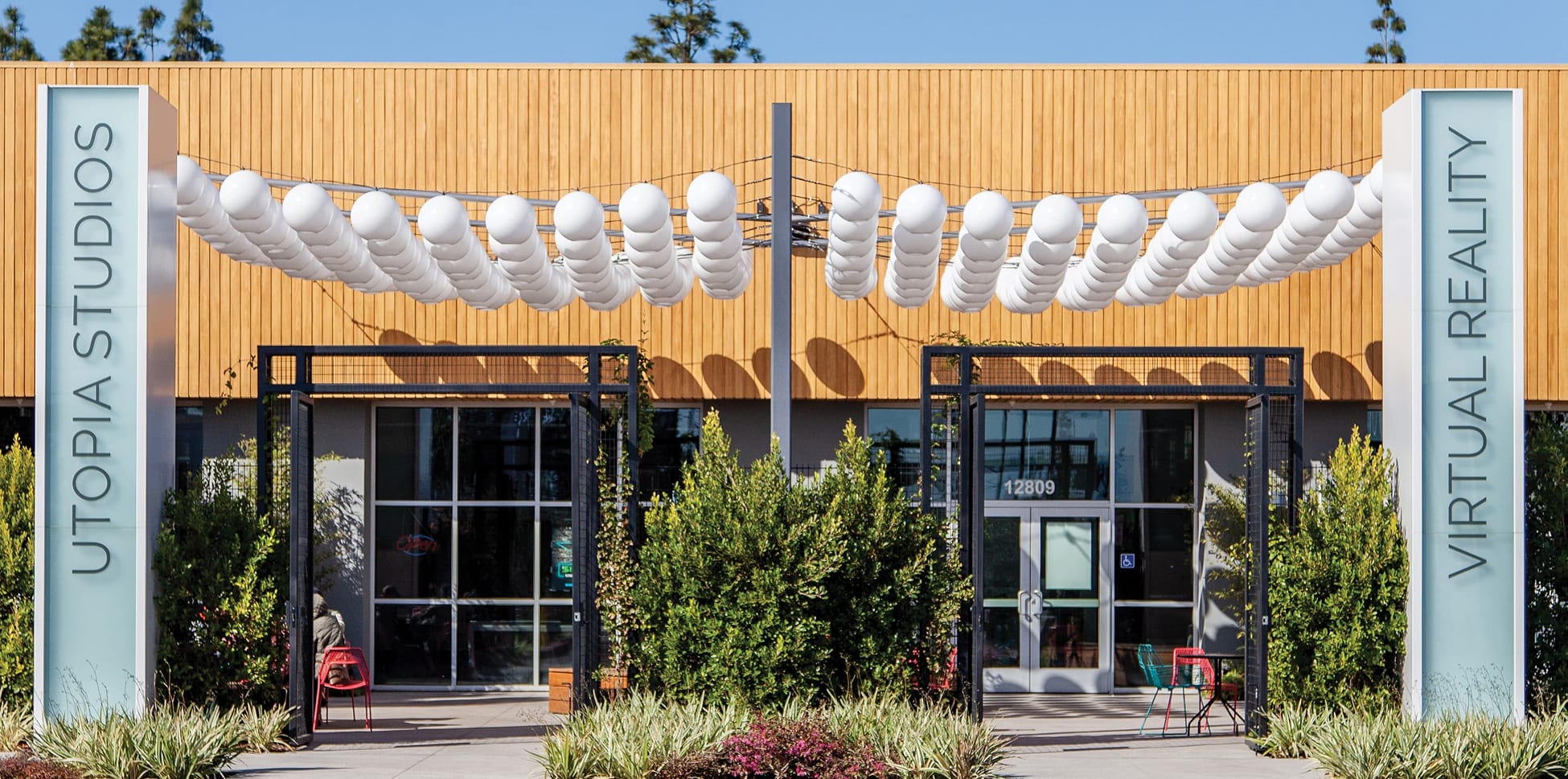 Daytime exterior of the Utopia Studios Virtual Reality tenant at Rodeo 39 Public Market in Stanton, CA, featuring a warm wood slat facade, black steel trellis entry framed by vertical illuminated signage, globe pendants strung overhead, and lush landscaping with colorful seating.