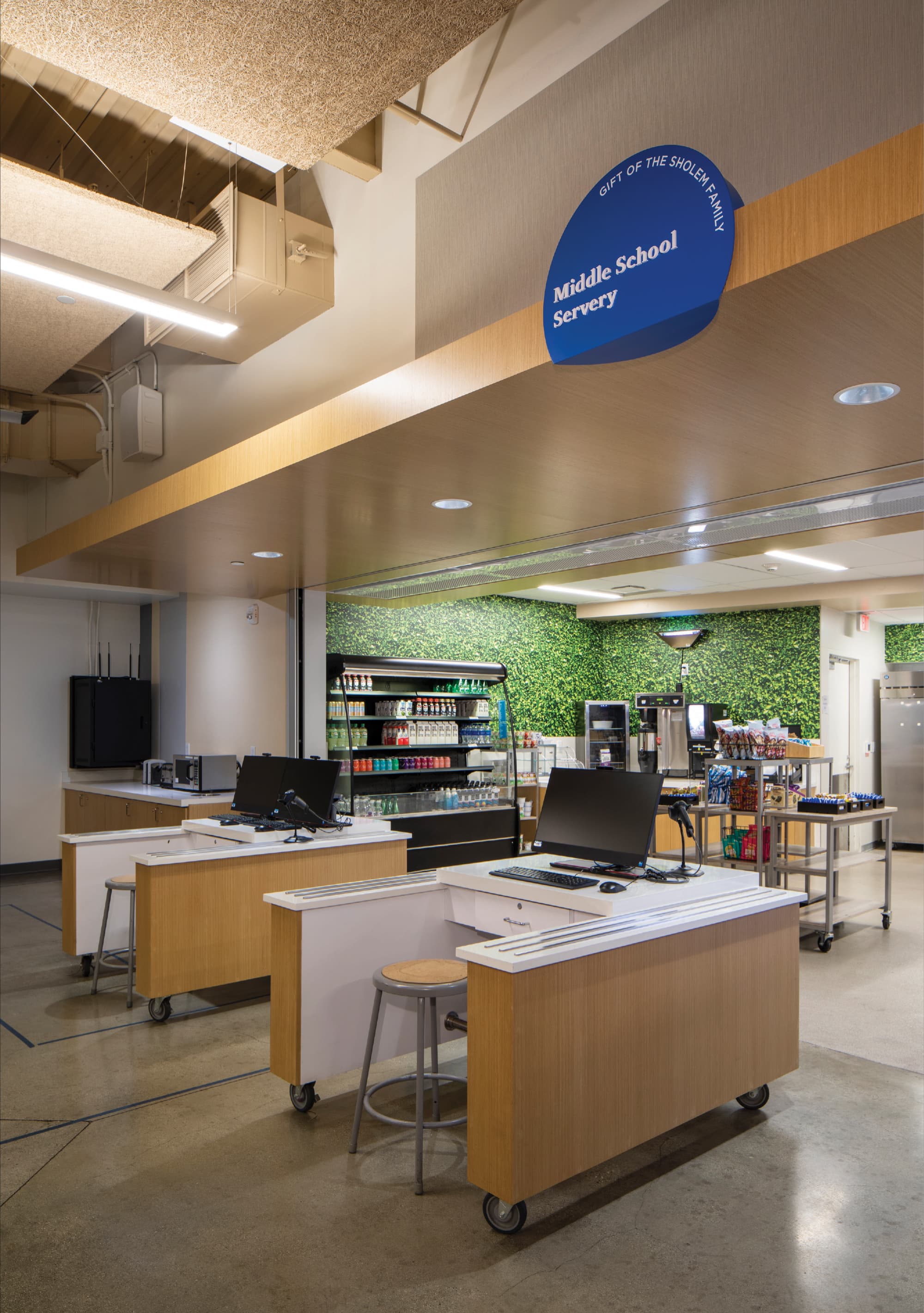 Interior of the Middle School Servery featuring a cobalt blue organic-shaped donor recognition and space identification sign reading "Middle School Servery — Gift of the Sholem Family" suspended from a wood-paneled ceiling beam above a cafeteria checkout counter, with a lush green living wall, refrigerated beverage display, and food service stations visible throughout — demonstrating RSM Design's donor recognition and specialty graphics and environmental graphic design services integrated into a student-centered space within the education market