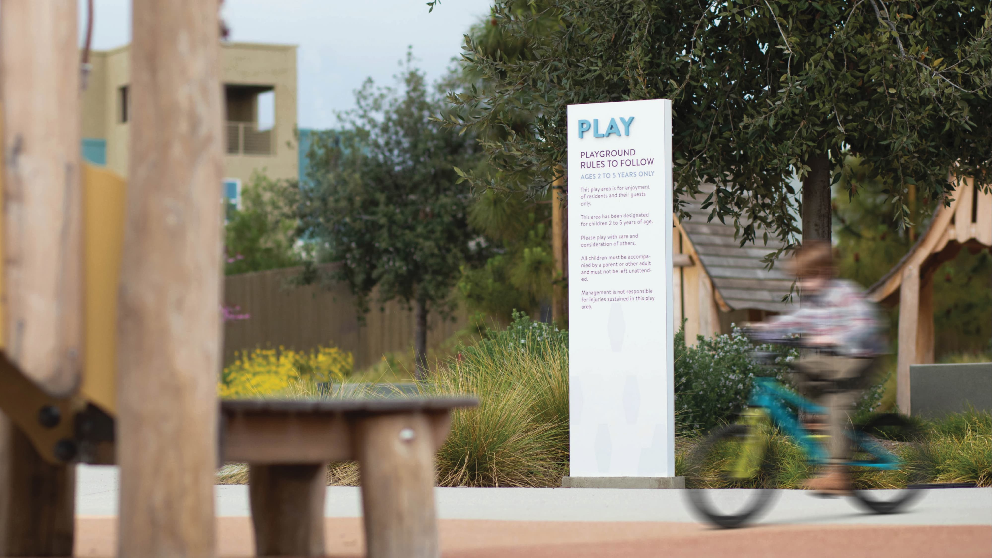 A white amenity identification and regulatory sign post reading "PLAY — Playground Rules to Follow, Ages 2 to 5 Years Only" positioned among native grasses and mature olive trees within a residential playground, with a child on a bicycle in motion in the background — illustrating RSM Design's wayfinding and signage design and placemaking services applied to the residential community market