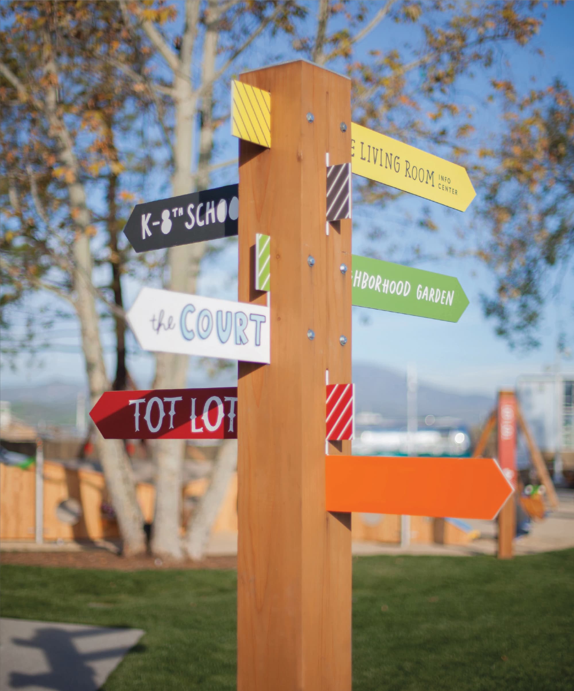 RSM Design pedestrian wayfinding signage and placemaking for FivePoint at Great Park Neighborhoods in Irvine, California, featuring a close-up of a wood post multi-directional arrow sign cluster with color-coded panels directing visitors to The Living Room Info Center, K-8th School, Neighborhood Garden, The Court, Tot Lot, and an orange directional arrow, each with unique typography and diagonal stripe accent details, set within an active community green space with autumn trees and playground structures visible in the background, demonstrating how environmental graphic design, specialty graphics, and wayfinding and signage design use color, personality, and playful typographic variety to create an intuitive, joyful, human-centered navigation system for residential community, education, and park markets.