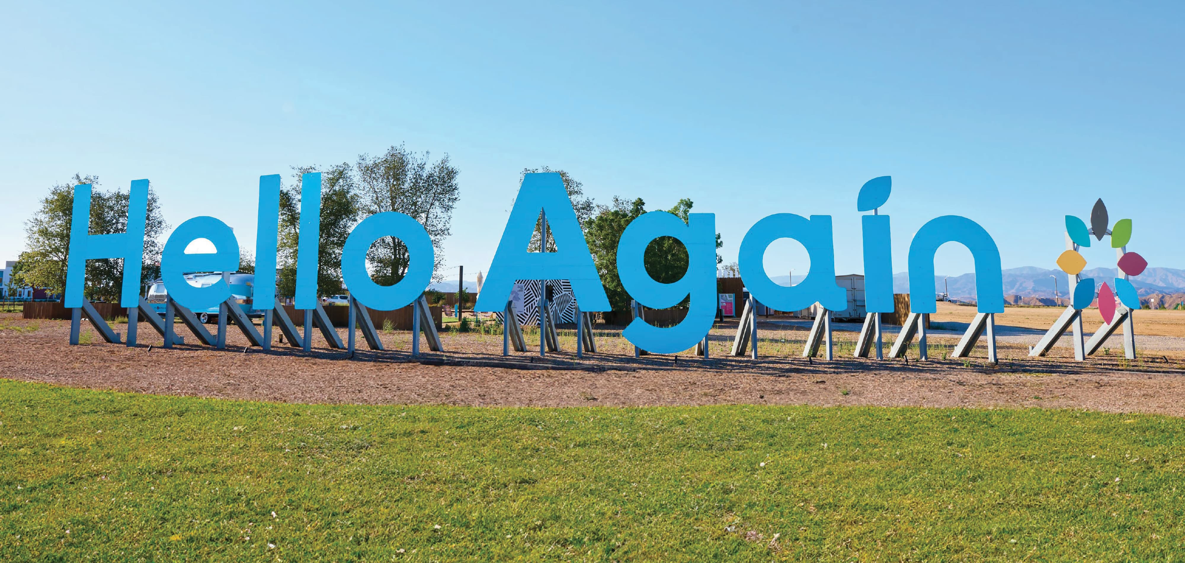 Large blue 'Hello Again' letters mounted on angled support beams over a gravel lot, with a colorful flower logo beside the text, trees and mountains in the background, and a grassy lawn in the foreground.
