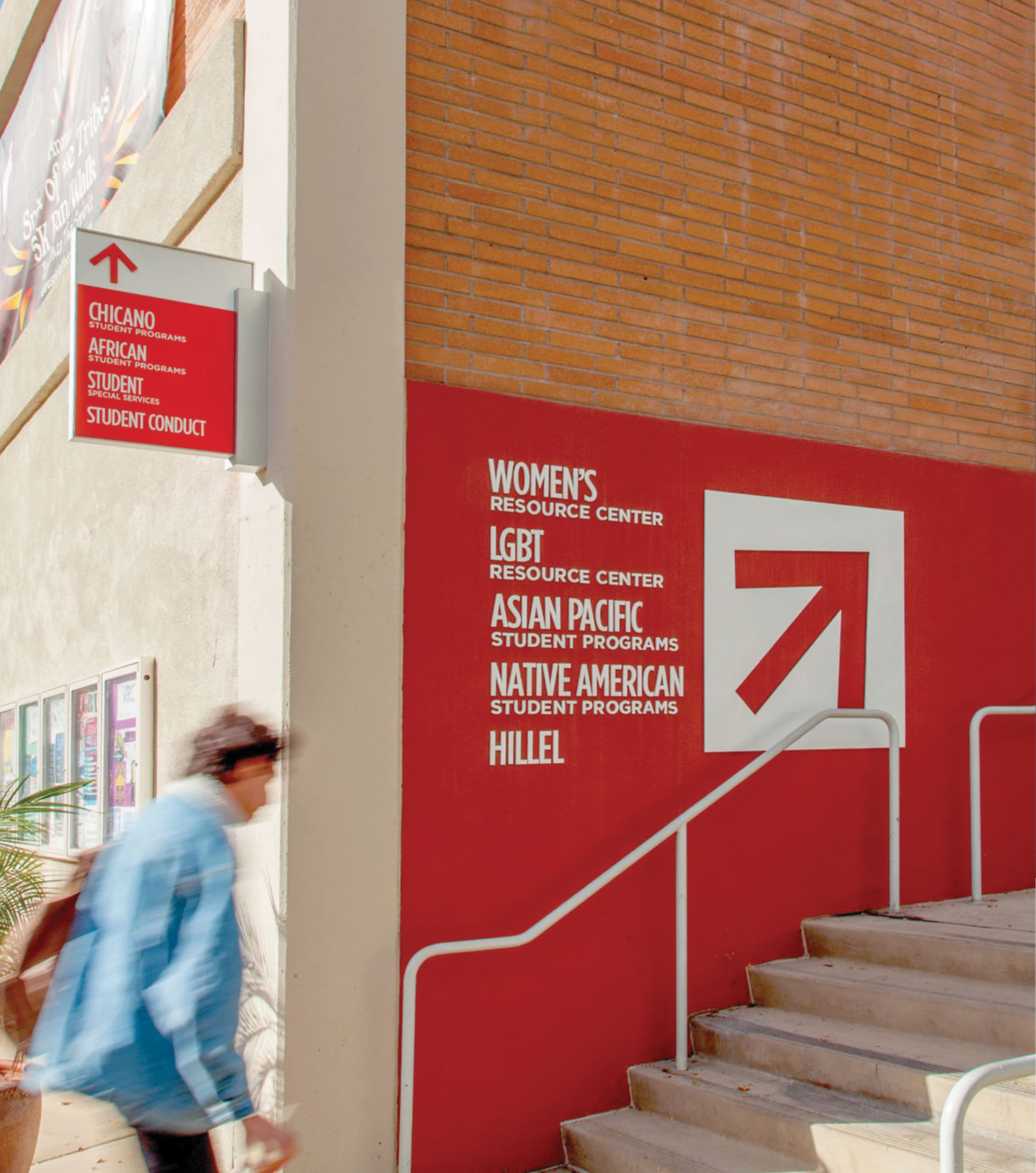 Red directional wayfinding sign listing student resource centers including Women's Resource Center, LGBT Resource Center, Asian Pacific Student Programs, Native American Student Programs, and Hillel, beside a stairway and a smaller wall-mounted sign for Chicano, African, and other student programs, as a person walks by.