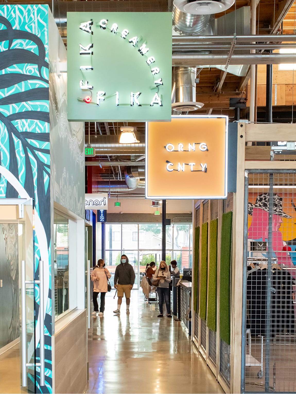 Interior corridor of Rodeo 39 Public Market with guests browsing vendor stalls, featuring illuminated hanging signs for Creamery Fika and ORNG CNTY, bold tropical and graphic murals, exposed ductwork, polished concrete floors, and moss wall accents beneath a wood beam ceiling.
