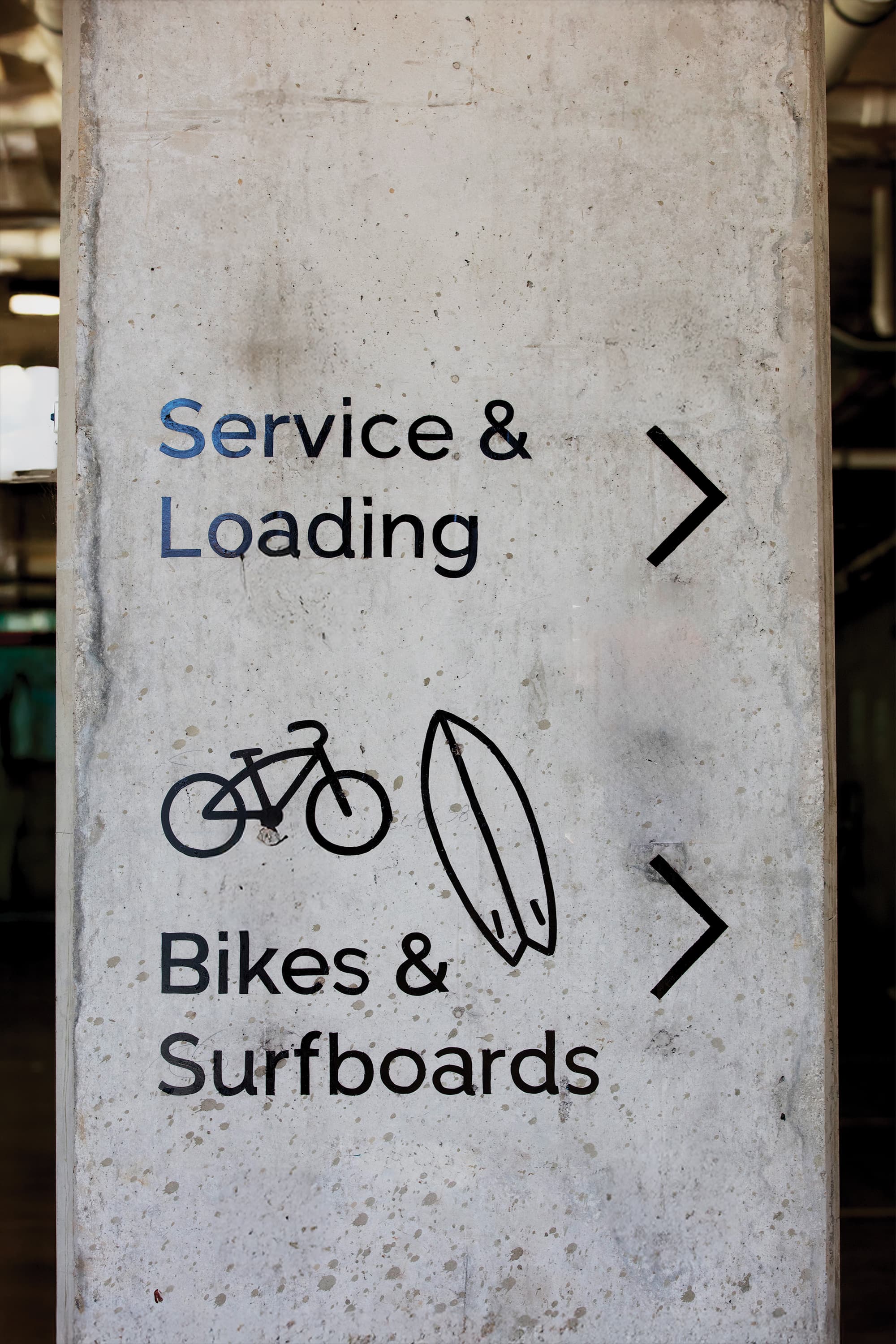 Concrete parking garage column with directional text reading 'Service & Loading' and 'Bikes & Surfboards,' each with a right-pointing arrow and a bicycle and surfboard icon.