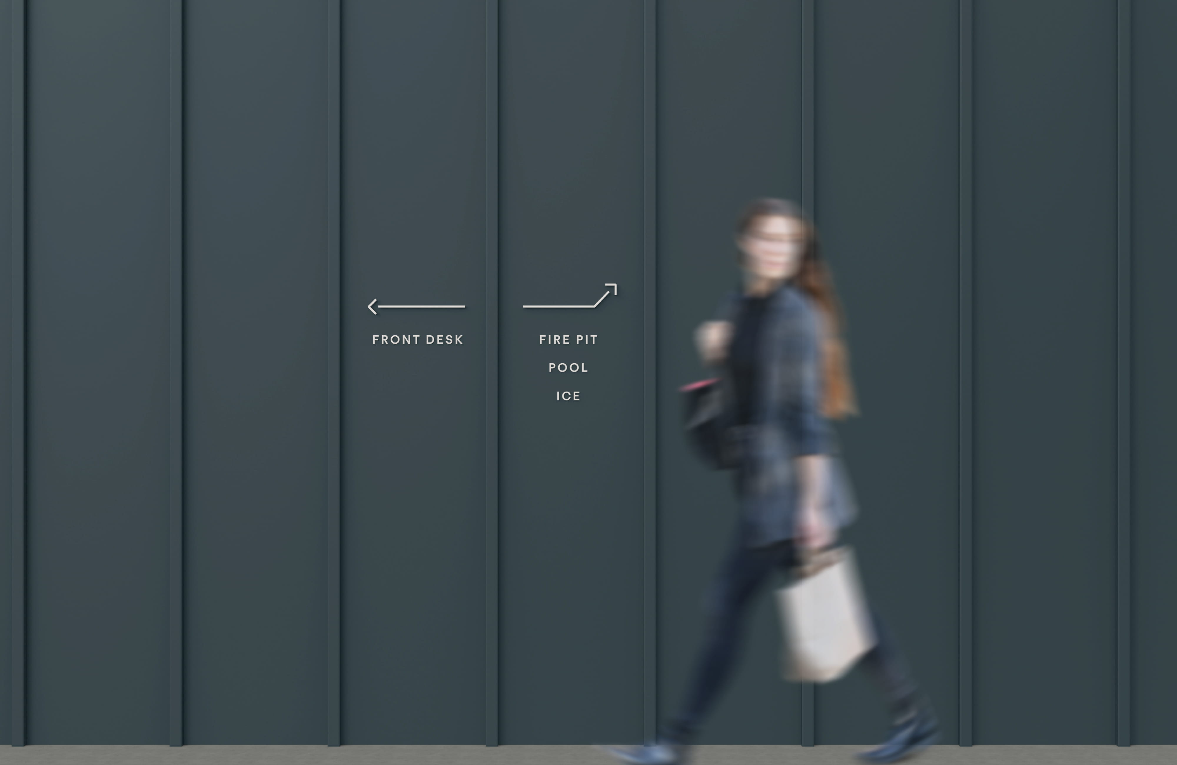 Guest walking past a dark teal exterior wall with minimalist directional wayfinding signage guiding visitors to the Front Desk, Fire Pit, Pool, and Ice at Trailborn Rocky Mountains Outpost, showcasing environmental graphic design and signage for a hospitality environment by RSM Design.