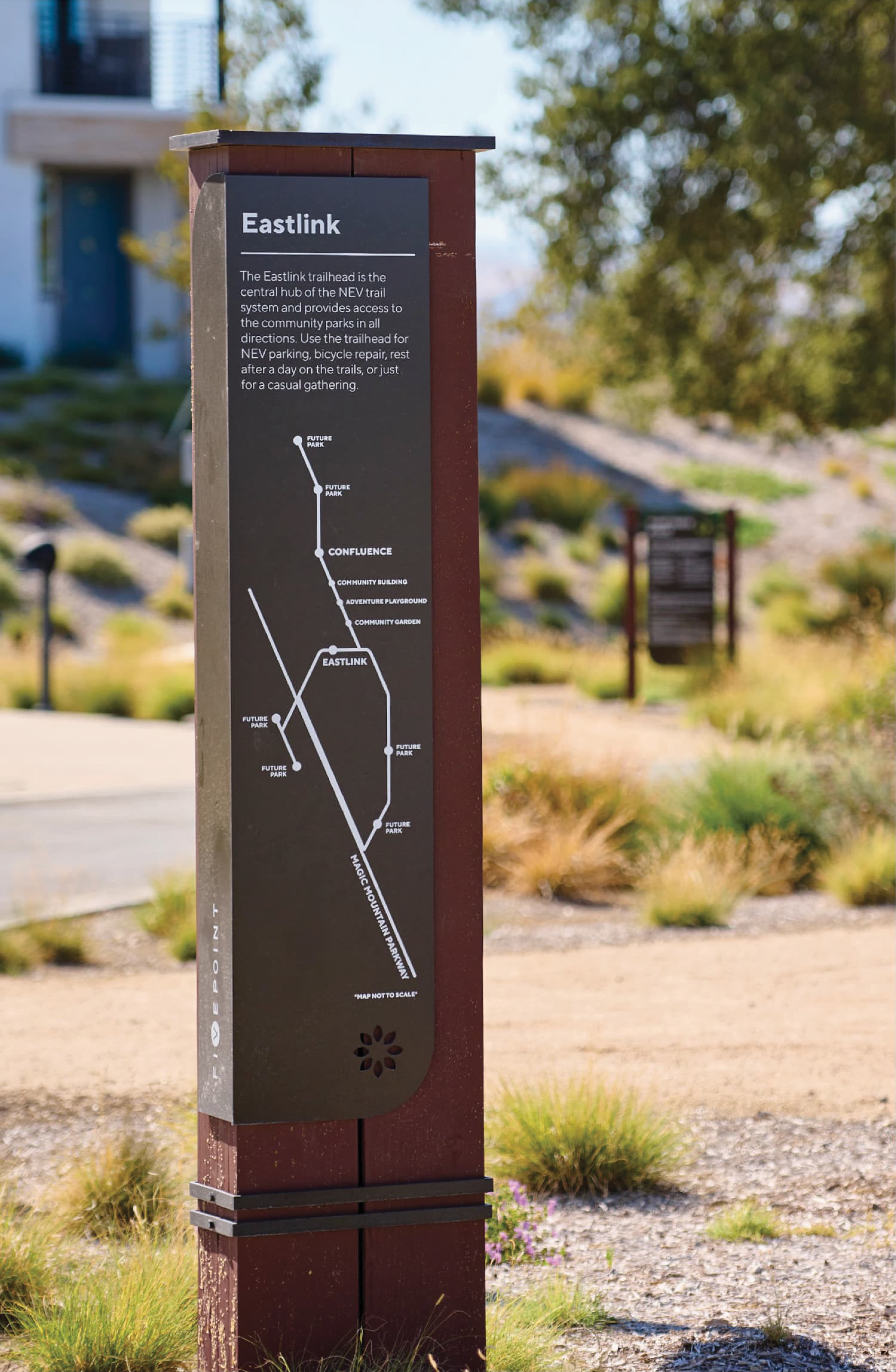 Eastlink trailhead wayfinding post with a transit-style map showing trail connections to Confluence, community parks, and future parks, set on a rusted metal post amid desert landscaping and another sign visible in the background.
