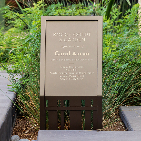 Tan sign reading 'Bocce Court & Garden, gifted in honor of Carol Aaron, with love and admiration by her children,' listing family names below, set in a landscaped bed of ornamental grasses.