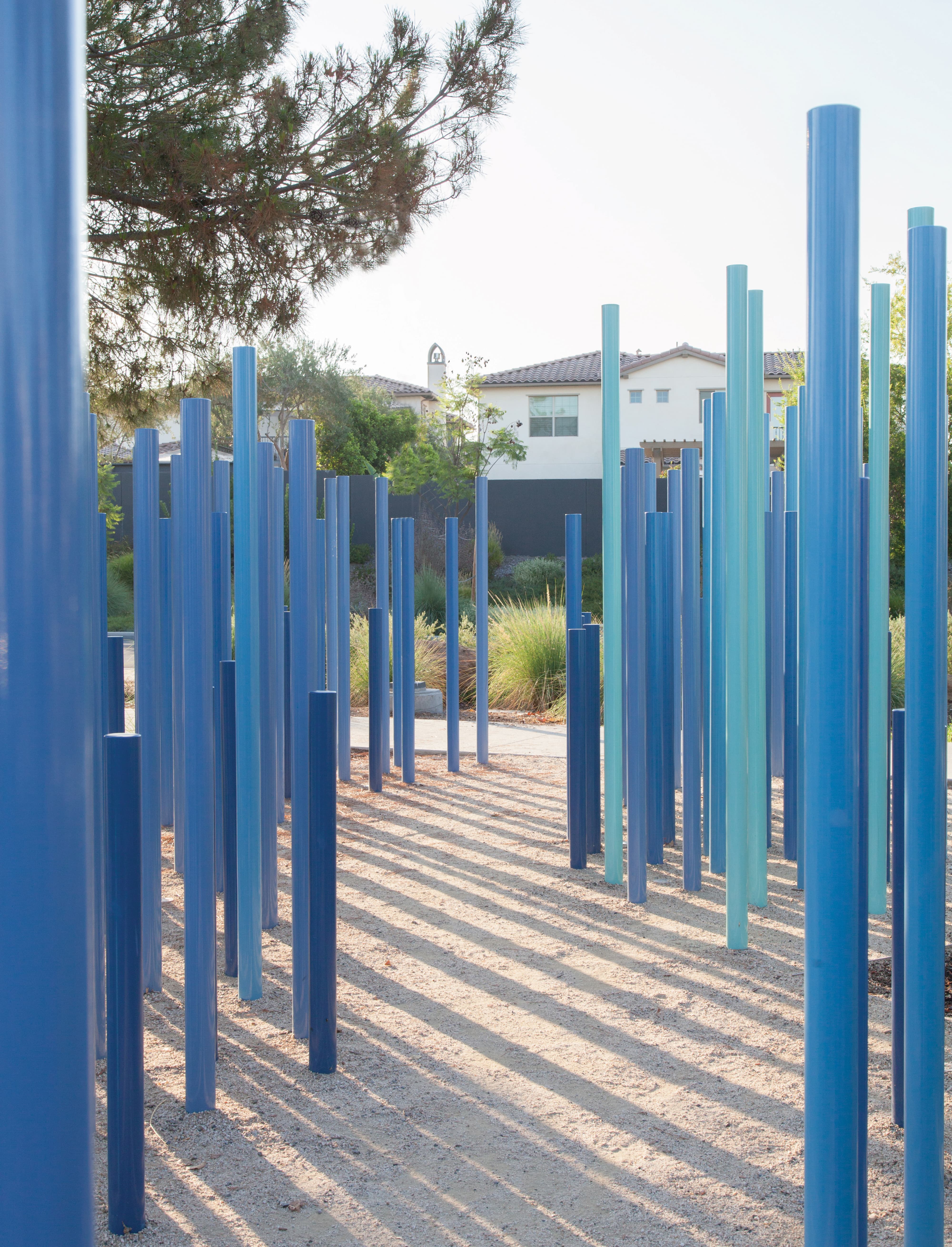 Art installation of tall vertical poles in varying shades of blue and teal, arranged in clusters over a sandy path, casting long shadows with a residential home visible in the background.