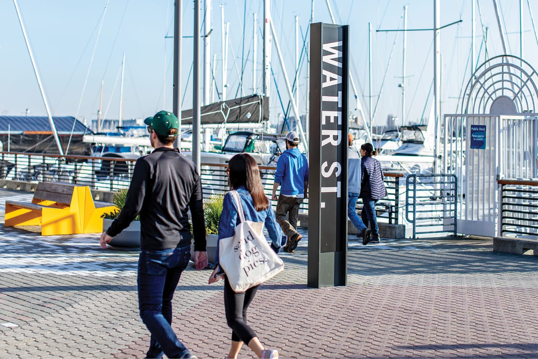 Waterfront promenade at Jack London Square in Oakland featuring a vertical “Water St.” wayfinding sign, marina with sailboats, public dock gate, and pedestrians walking along the harbor boardwalk on a sunny day.