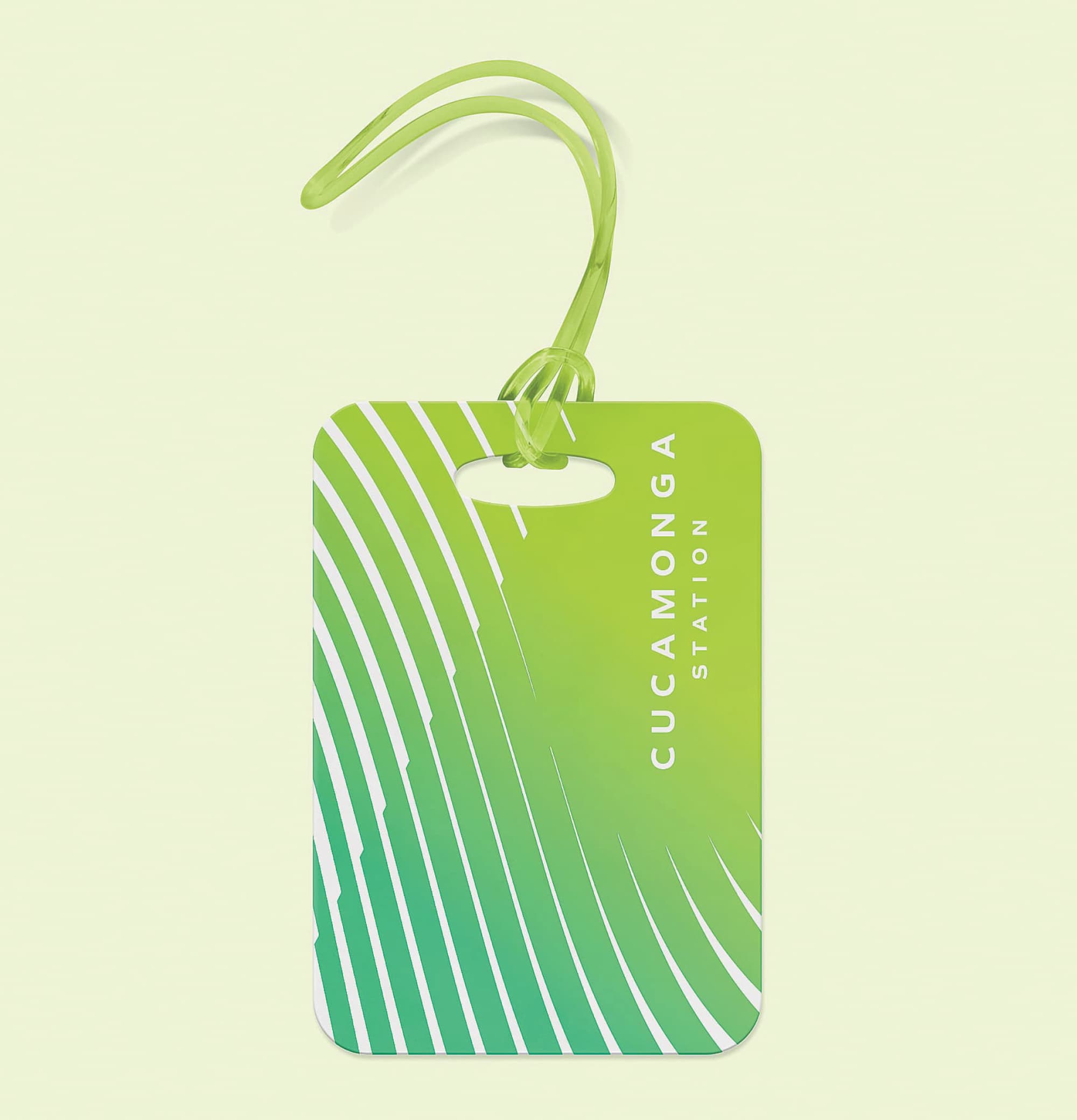 A Cucamonga Station branded luggage tag featuring a teal-to-lime green gradient background with sweeping white diagonal line graphics and the vertical "Cucamonga Station" wordmark in white, looped with a matching lime green cord — illustrating RSM Design's branding and visual identity and print collateral services applied across branded environment touchpoints within the transit market