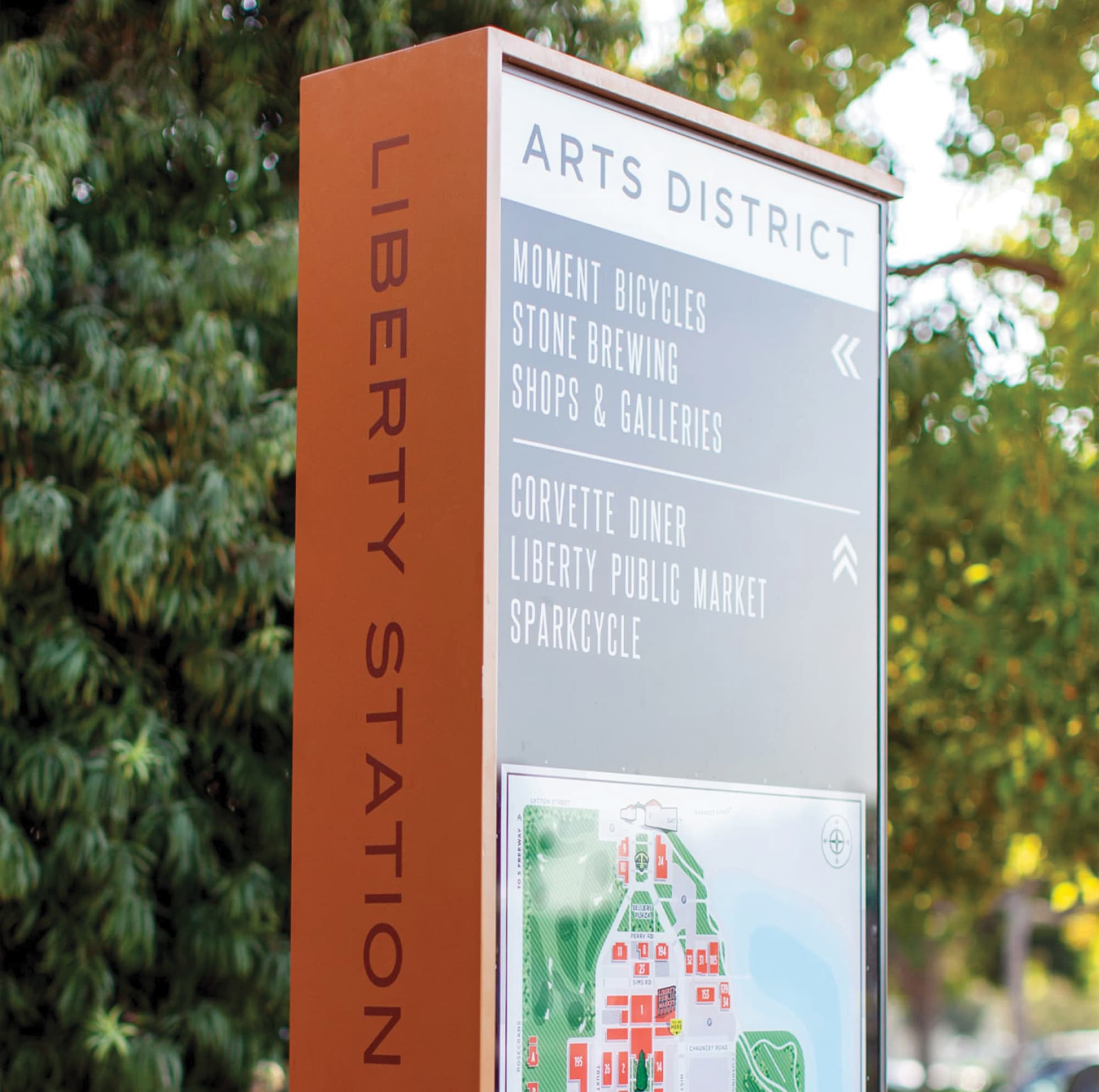 Orange 'Liberty Station' wayfinding pylon reading 'Arts District' with directional listings for Moment Bicycles, Stone Brewing, Shops & Galleries, Corvette Diner, Liberty Public Market, and SparkCycle, above a color-coded site map, set against green trees.