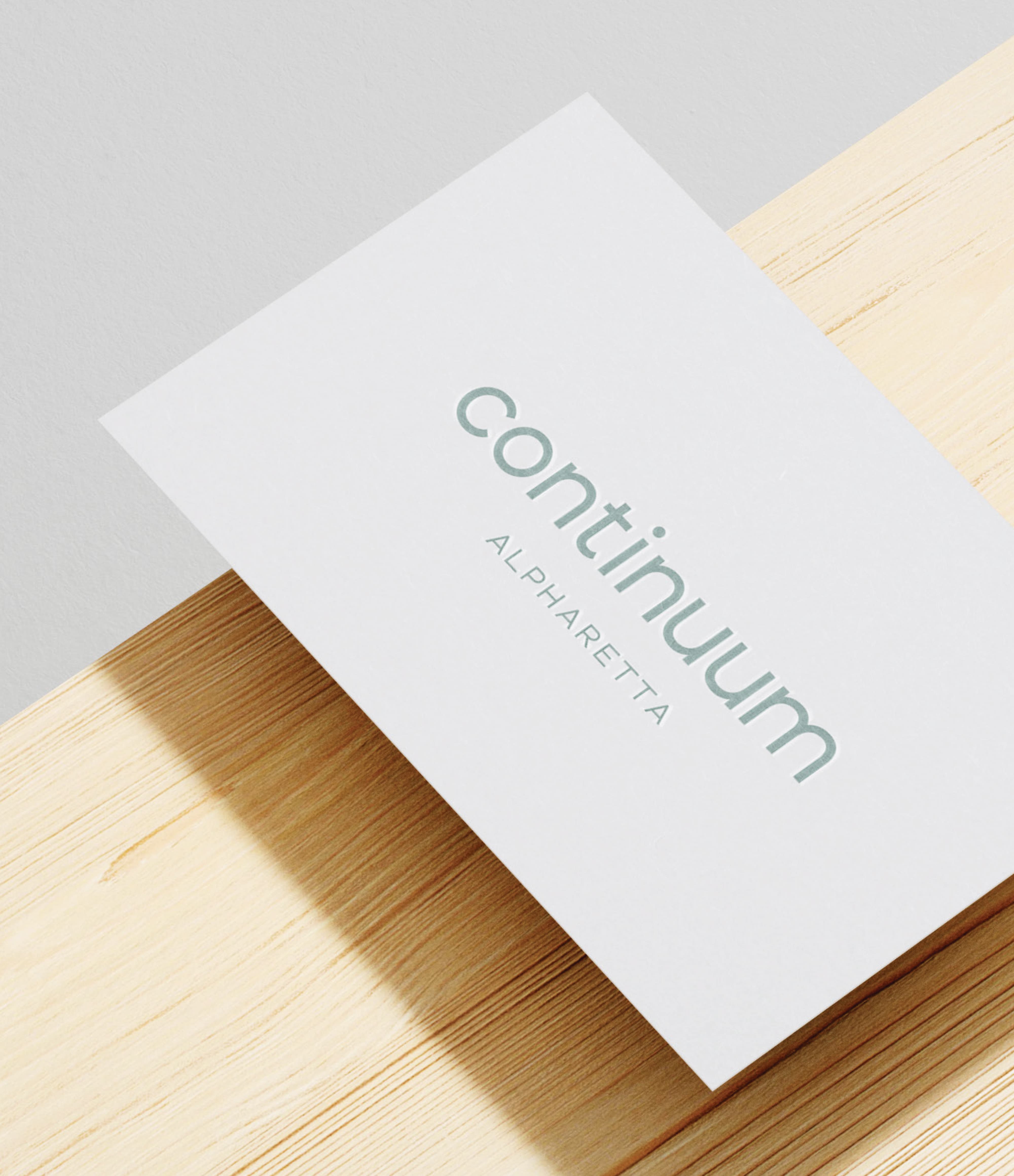 RSM Design print collateral and visual identity design for Continuum Alpharetta, featuring a close-up of a sage green logo letterpress or tone-on-tone printed business card resting on a light wood surface, showcasing the refined typographic character and tactile print quality of the brand identity system developed as part of a comprehensive branding, visual identity, and print and digital collateral program for a mixed-use workplace, health and wellness, and residential community development.