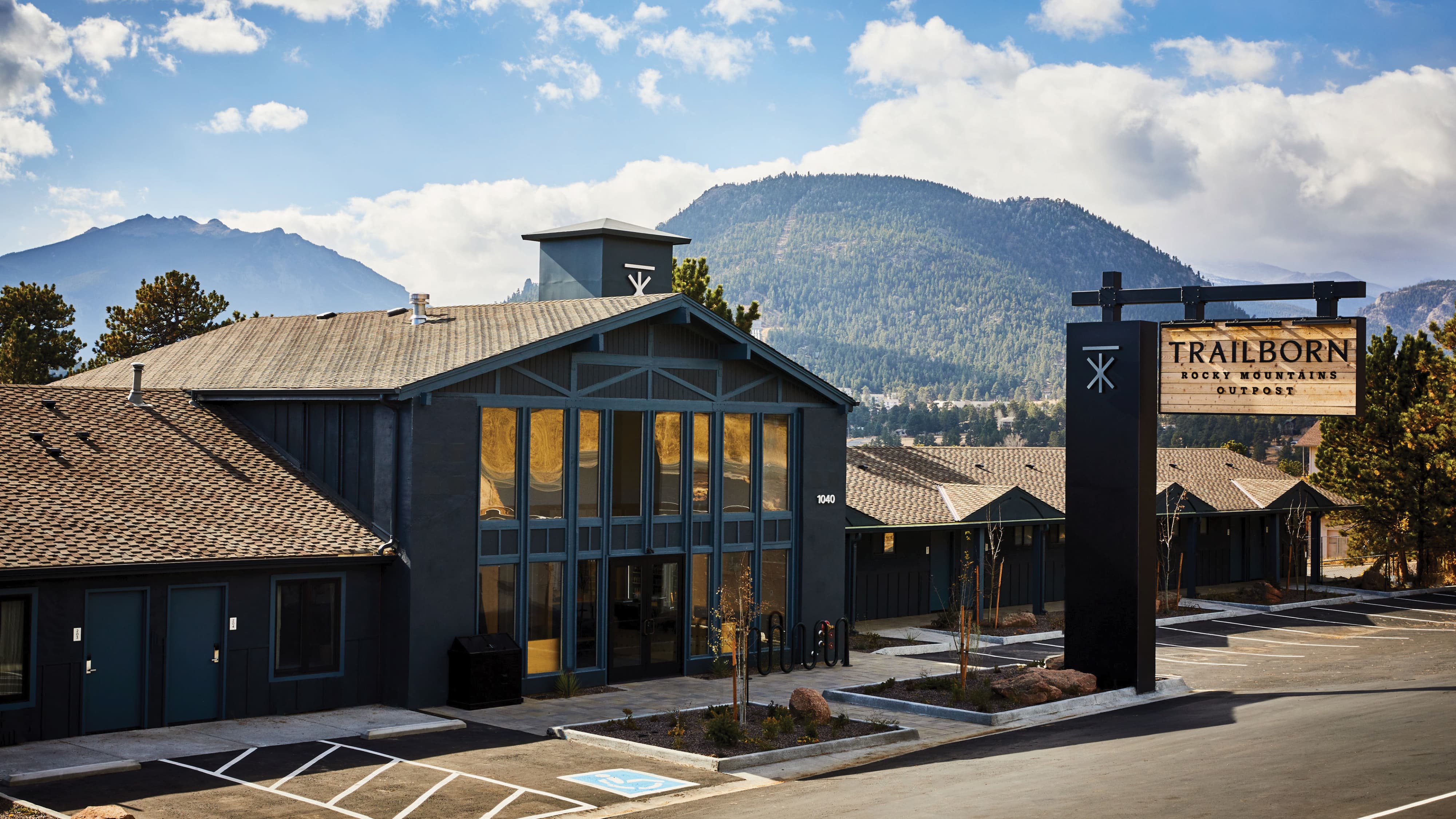 Exterior of Trailborn Rocky Mountains Outpost hotel featuring a tall black monument sign with wood panel inset and brand icon against a dramatic Rocky Mountain backdrop, showcasing environmental graphic design and signage for a hospitality environment by RSM Design.