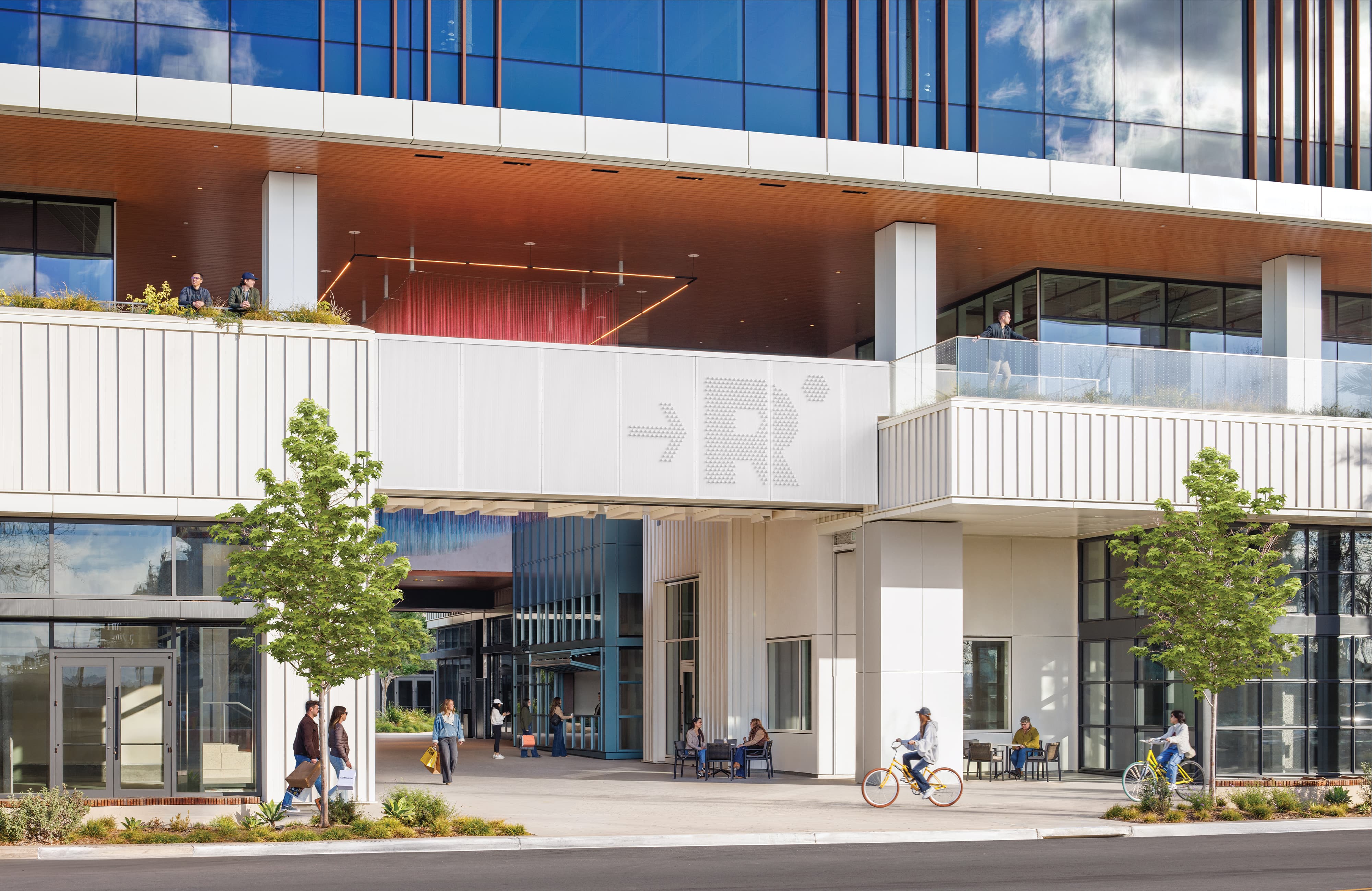 Modern mixed-use building exterior with a perforated metal signage panel featuring an arrow and stylized lettering above a covered walkway, where people walk, sit at outdoor tables, and ride bicycles past ground-floor retail storefronts.