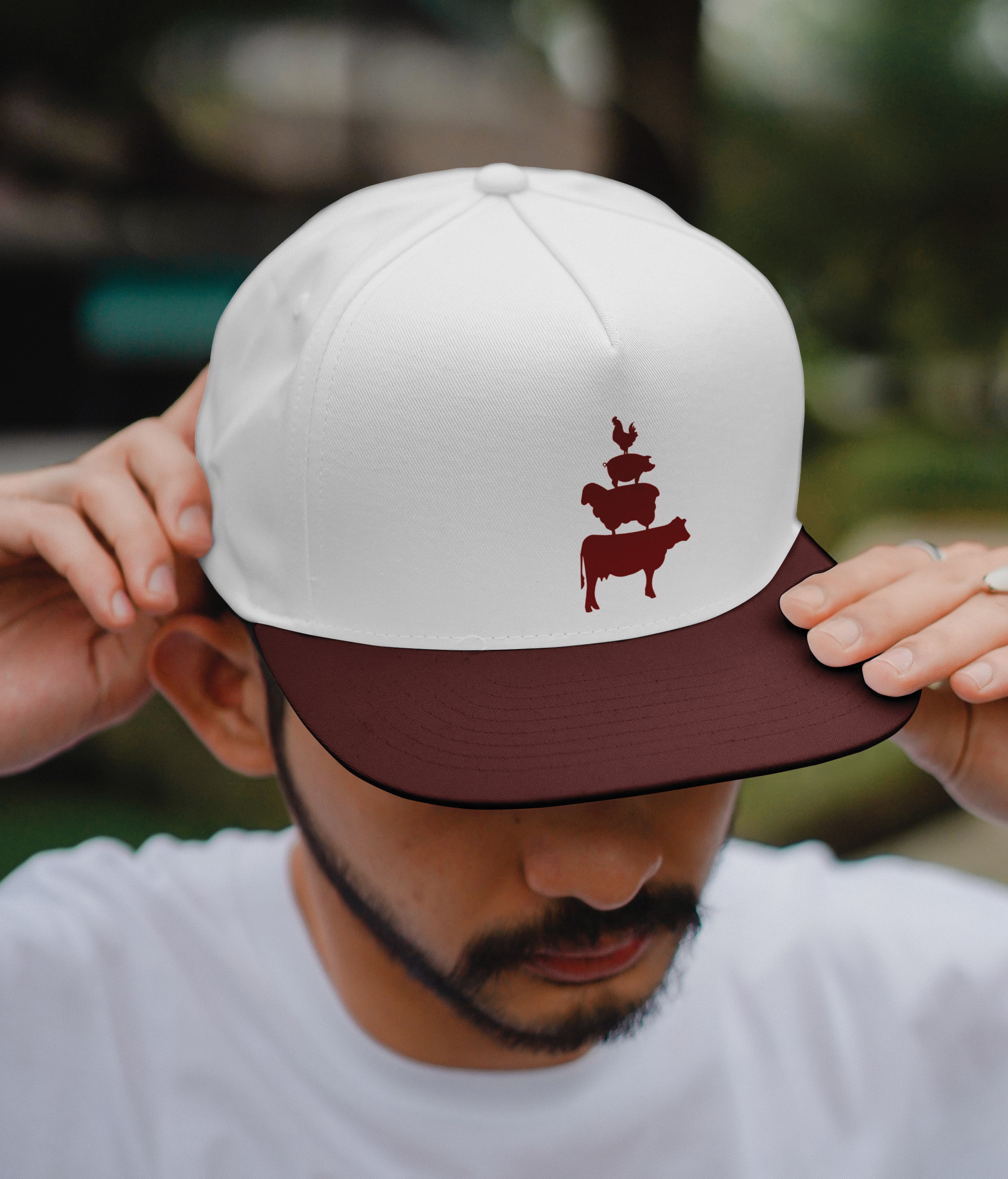 White and burgundy snapback cap embroidered with River Street Marketplace's stacked farm animal brand icon, showcasing specialty graphics and visual identity design for a mixed-use waterfront retail environment by RSM Design.
