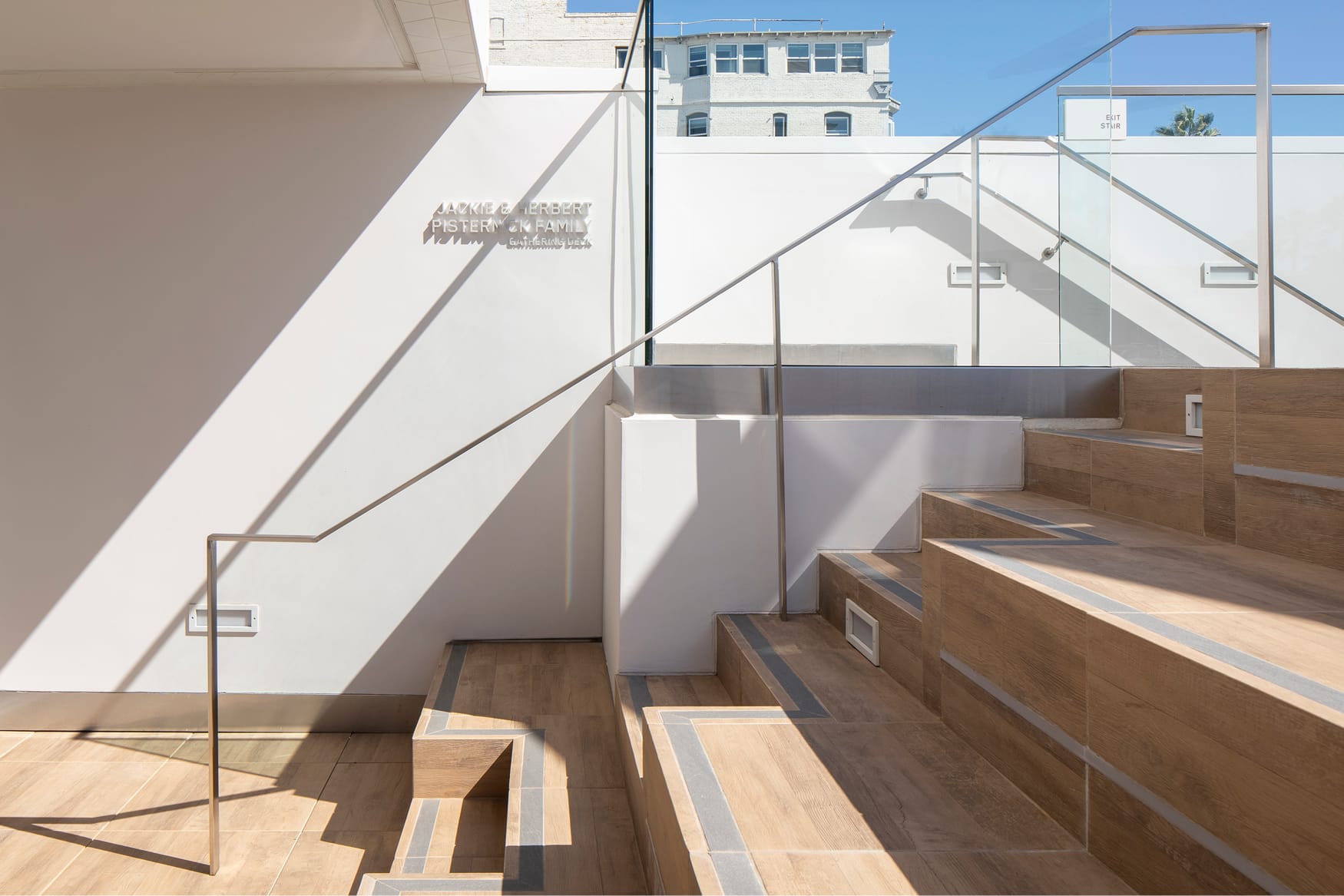 Sunlit outdoor rooftop terrace and staircase of the Jackie and Herbert Pisternick Family Gathering Deck featuring white-on-white dimensional donor recognition lettering, warm wood-look porcelain tile steps with integrated step lighting, frameless glass panel railings with brushed steel handrails, a built-in bench, and open views of the surrounding Venice Beach Los Angeles urban skyline under a bright blue sky