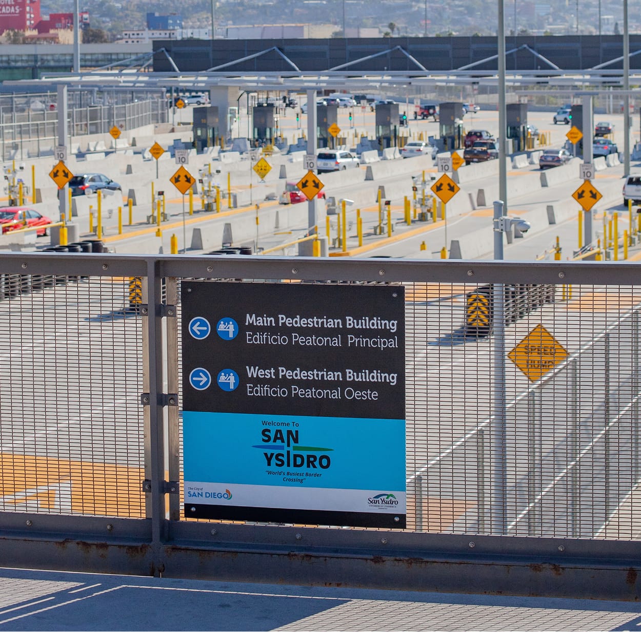Bilingual wayfinding sign mounted on a mesh pedestrian bridge railing, directing to the Main Pedestrian Building and West Pedestrian Building with a 'Welcome to San Ysidro' banner below, overlooking rows of vehicle inspection booths and lanes of cars at the border crossing.