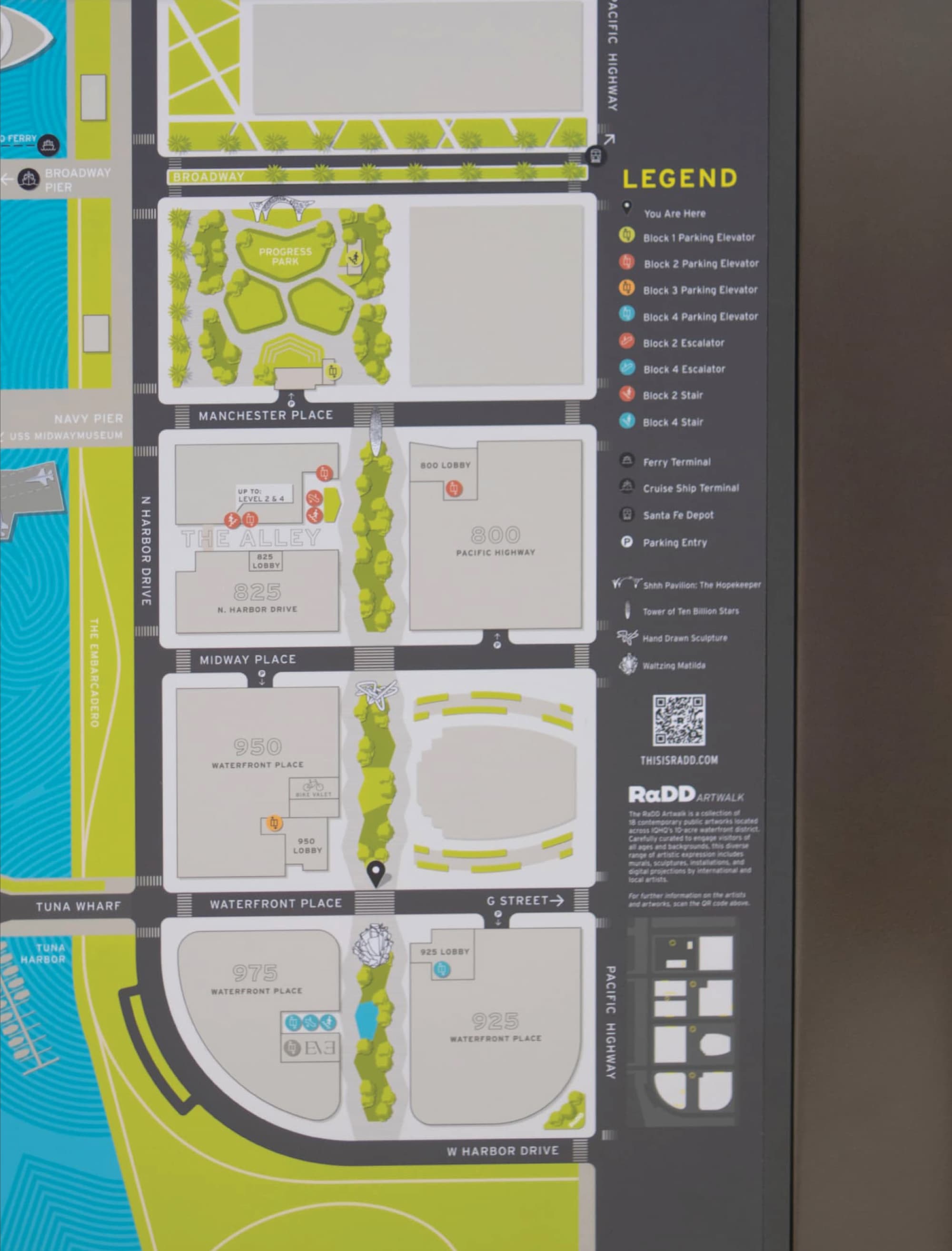 Detailed campus map sign for RαDD's waterfront district in San Diego, showing labeled buildings including The Alley, 800, 825, 950, 975, and 925 Waterfront Place, Progress Park, and a color-coded legend for parking elevators, escalators, stairs, ferry and cruise terminals, and public artworks, with a QR code linking to thisisradd.com.
