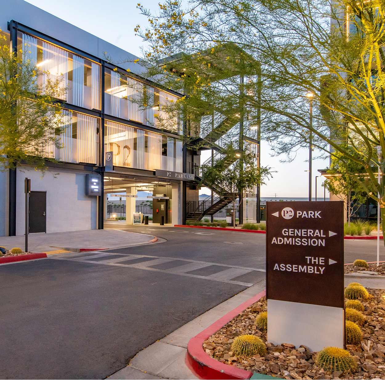 Exterior view of the P2 parking structure at Uncommons at dusk, with a dark wayfinding sign in the foreground directing visitors left to P2 Park, right to General Admission, and right to The Assembly. The multi-level parking structure features illuminated sheer screen facades and an exposed exterior staircase. Golden barrel cacti and desert trees frame the drive aisle and signage.