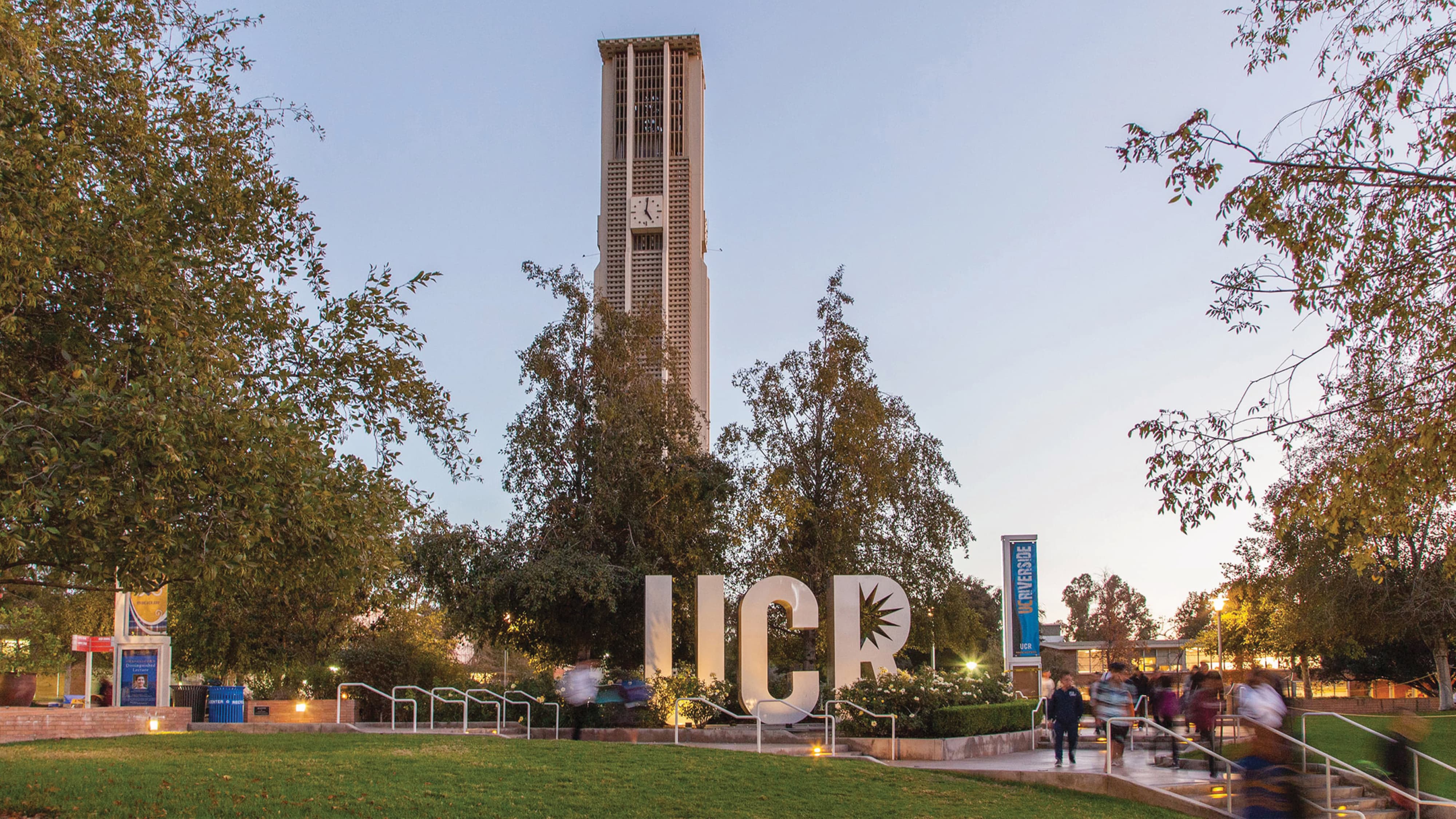 Twilight view of the UC Riverside Bell Tower rising above trees, with a large 'UCR' letter sculpture and 'UC Riverside' banner in the foreground, as students walk down illuminated steps.