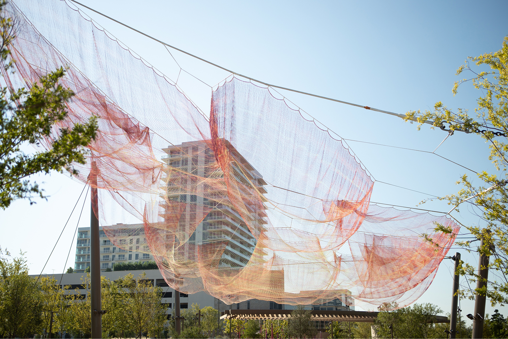 Large-scale suspended mesh net public art installation in warm pink and orange tones floating above tree canopy at Kaleidoscope Park with mixed-use residential tower in background, Frisco, Texas