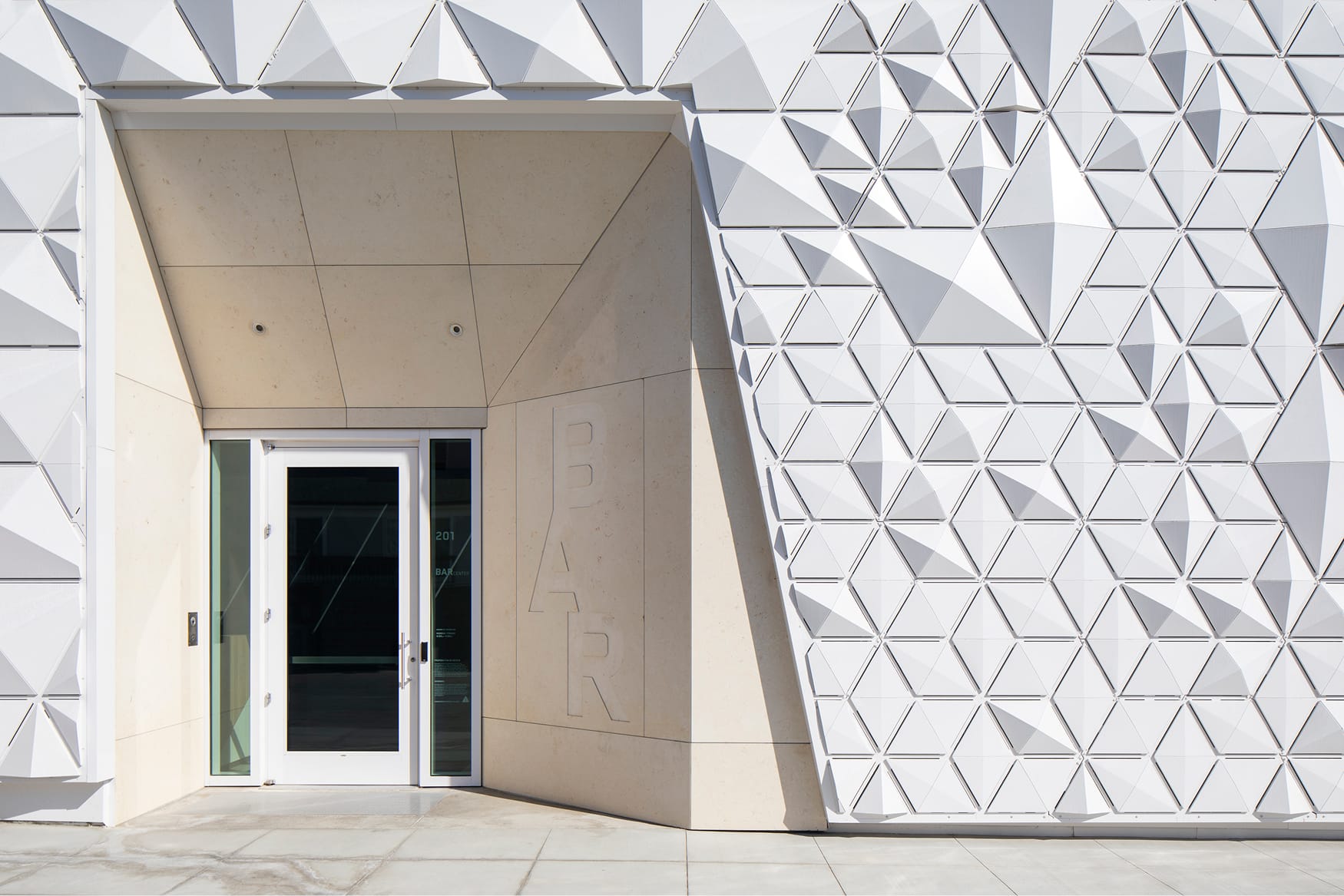 Exterior entrance of a contemporary architectural building featuring a dramatic three-dimensional white triangular geometric facade, smooth concrete canopy overhang, glass double doors, and embossed BAR lettering on the wall