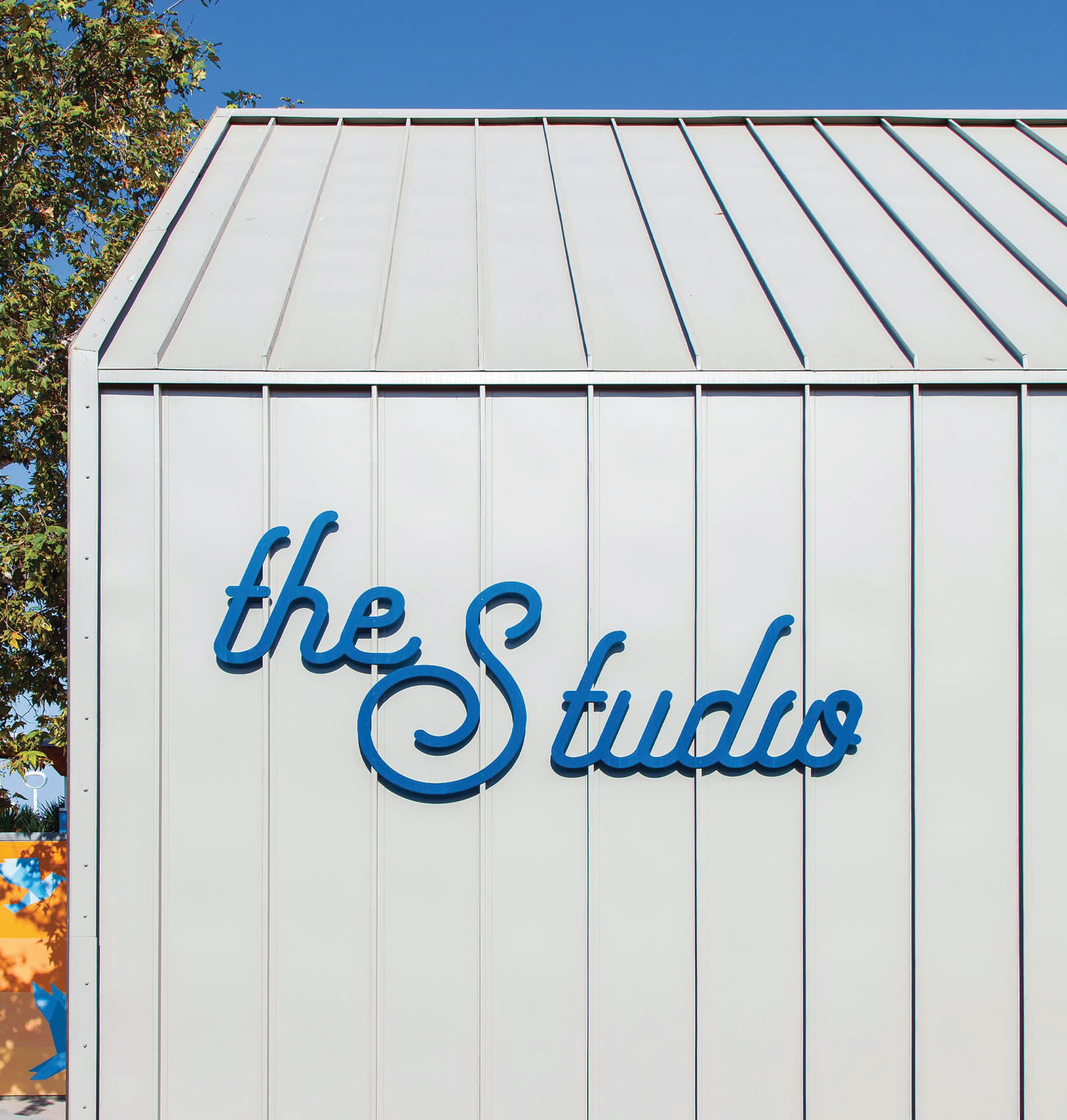 Dimensional blue script lettering reading "the Studio" mounted on a metal-clad exterior wall at a residential community amenity, showcasing branded environment and experiential graphic design by RSM Design.