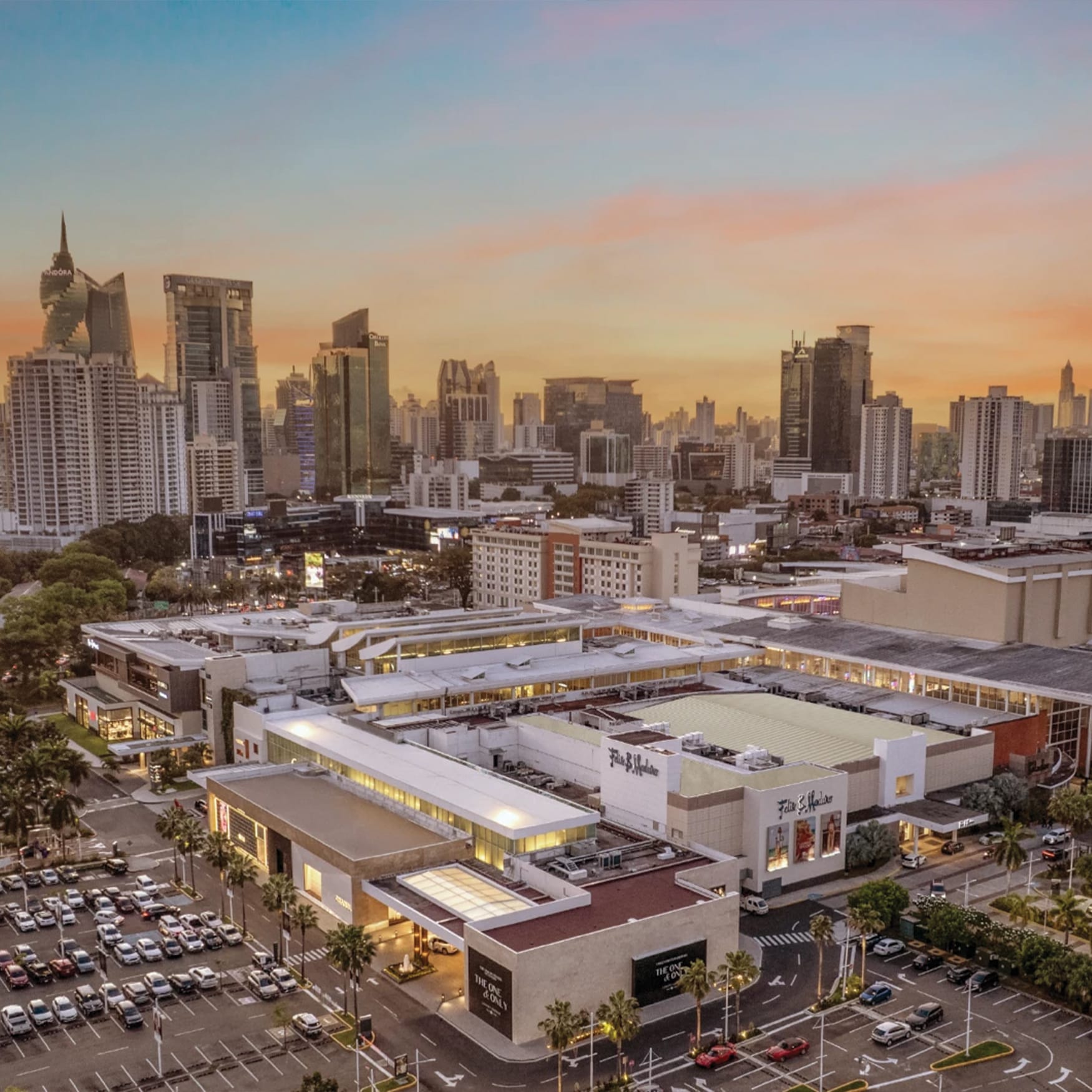 Aerial view of a modern urban shopping complex at sunset with palm-lined streets, parking areas, and illuminated storefronts, set against a dense city skyline with high-rise buildings in the background.
