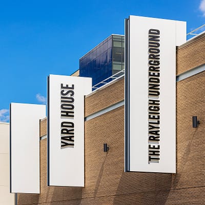 Exterior tenant identification blade signs for Yard House and The Rayleigh Underground mounted on a brick building facade at Toyota Music Factory mixed-use entertainment district in Irving, Texas — RSM Design wayfinding and signage design, master sign programs, and branded environments for mixed-use, food and beverage, and retail destinations.