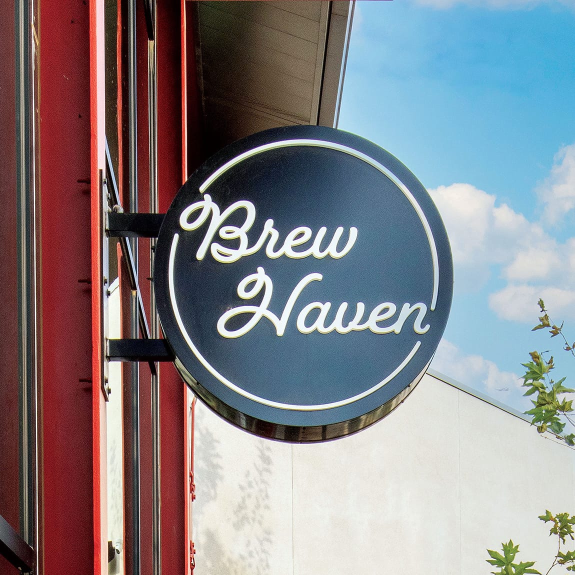 Circular black blade sign reading 'Brew Haven' in white script, mounted on a red building column against a blue sky.