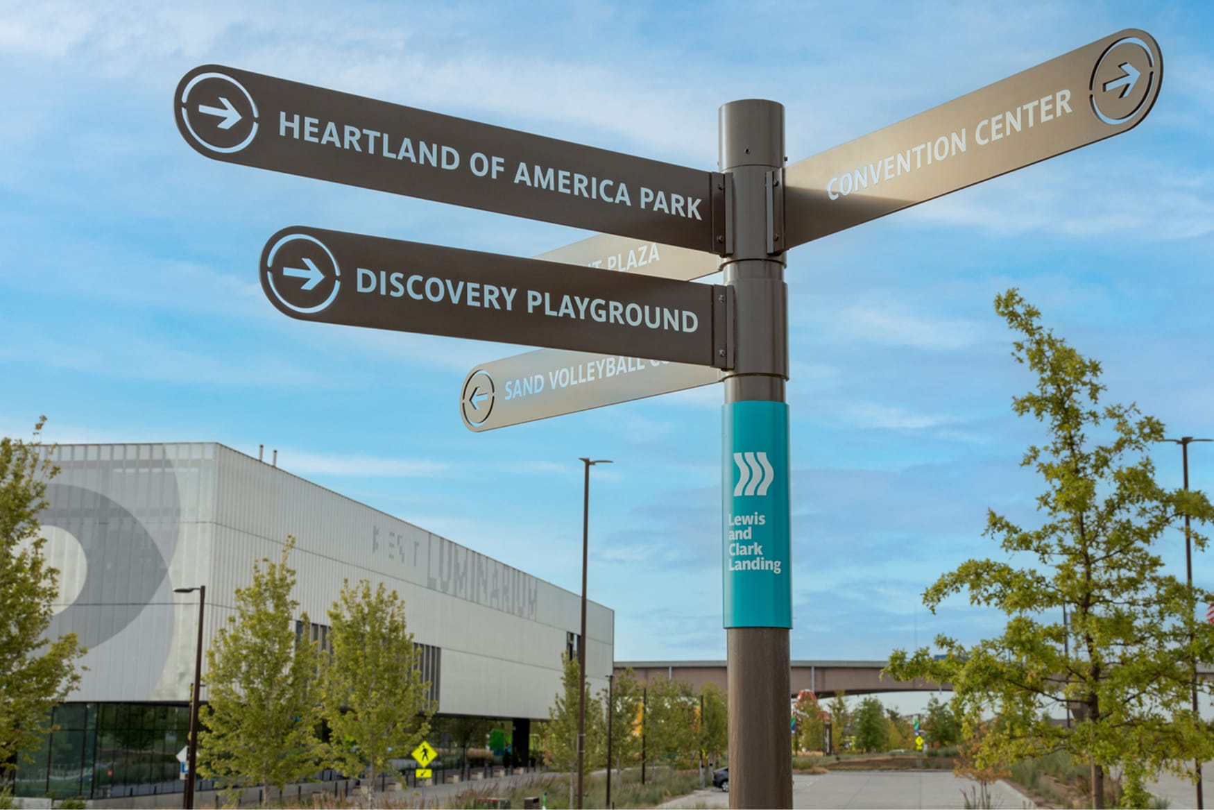 Lewis and Clark Landing wayfinding sign in Omaha, Nebraska, directing visitors to Heartland of America Park, Discovery Playground, Convention Center, and Sand Volleyball Courts, with the Fest Luminarium building visible in the background