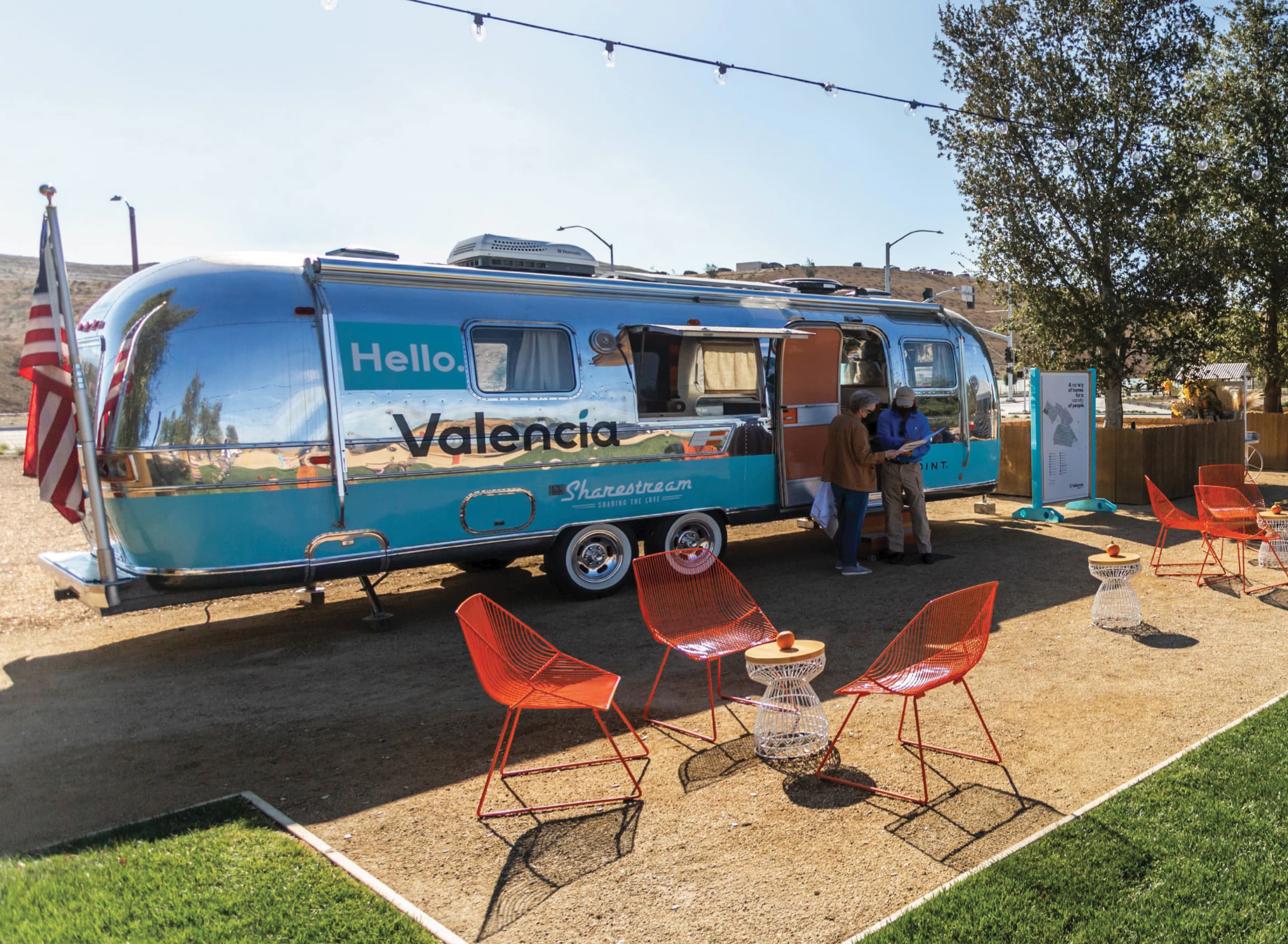 Vintage silver Airstream trailer branded 'Hello, Valencia' and 'Sharestream,' with two visitors chatting at its open door, surrounded by red wire chairs, string lights, an American flag, and a wayfinding map sign under a clear sky