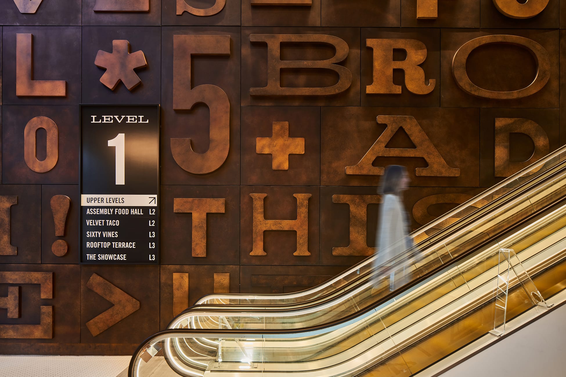 Level 1 wayfinding sign at Fifth + Broadway set against a large-scale typographic wall, guiding visitors to key destinations while reinforcing the district’s bold visual identity.