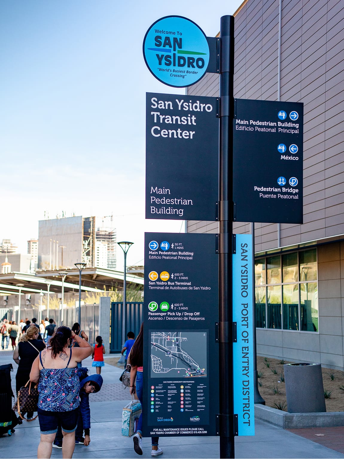 Comprehensive bilingual wayfinding sign at the San Ysidro Transit Center with directions to the Main Pedestrian Building, Mexico, Pedestrian Bridge, bus terminal, and passenger pick-up, plus an area map and 'San Ysidro Port of Entry District' label, as pedestrians walk past a transit building.