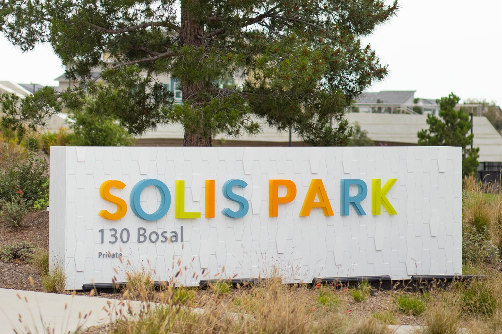 Solis Park neighborhood entrance monument sign with multicolor dimensional letters in orange, teal, and yellow-green mounted on a white textured mosaic tile wall at 130 Bosal Private in Irvine, California, designed by RSM Design