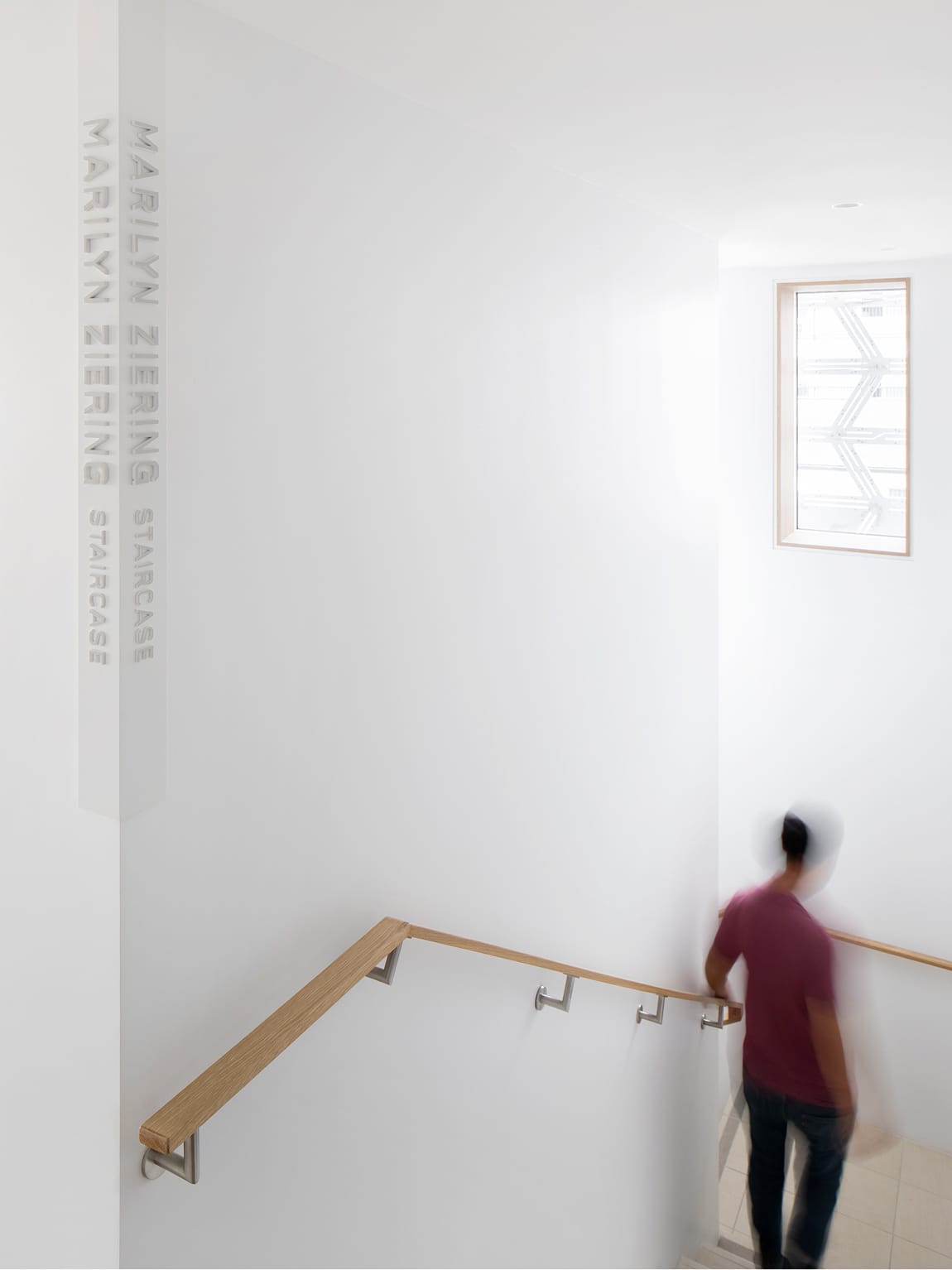 Bright minimalist interior stairwell of the Marilyn Ziering Staircase featuring vertical white-on-white dimensional donor recognition lettering on the wall, warm wood handrail with brushed steel brackets, a skylight window reflecting the geometric building facade, and a motion-blurred person ascending the stairs