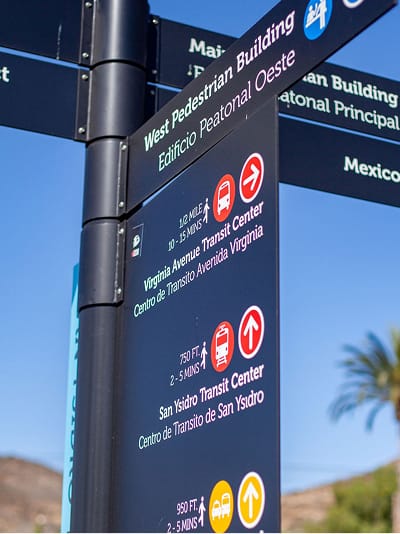 Cluster of bilingual directional signs at San Ysidro pointing to the West Pedestrian Building, Main Pedestrian Building, Mexico, Virginia Avenue Transit Center, and San Ysidro Transit Center, each listing walking distance and time, against a blue sky with a palm tree and hills.