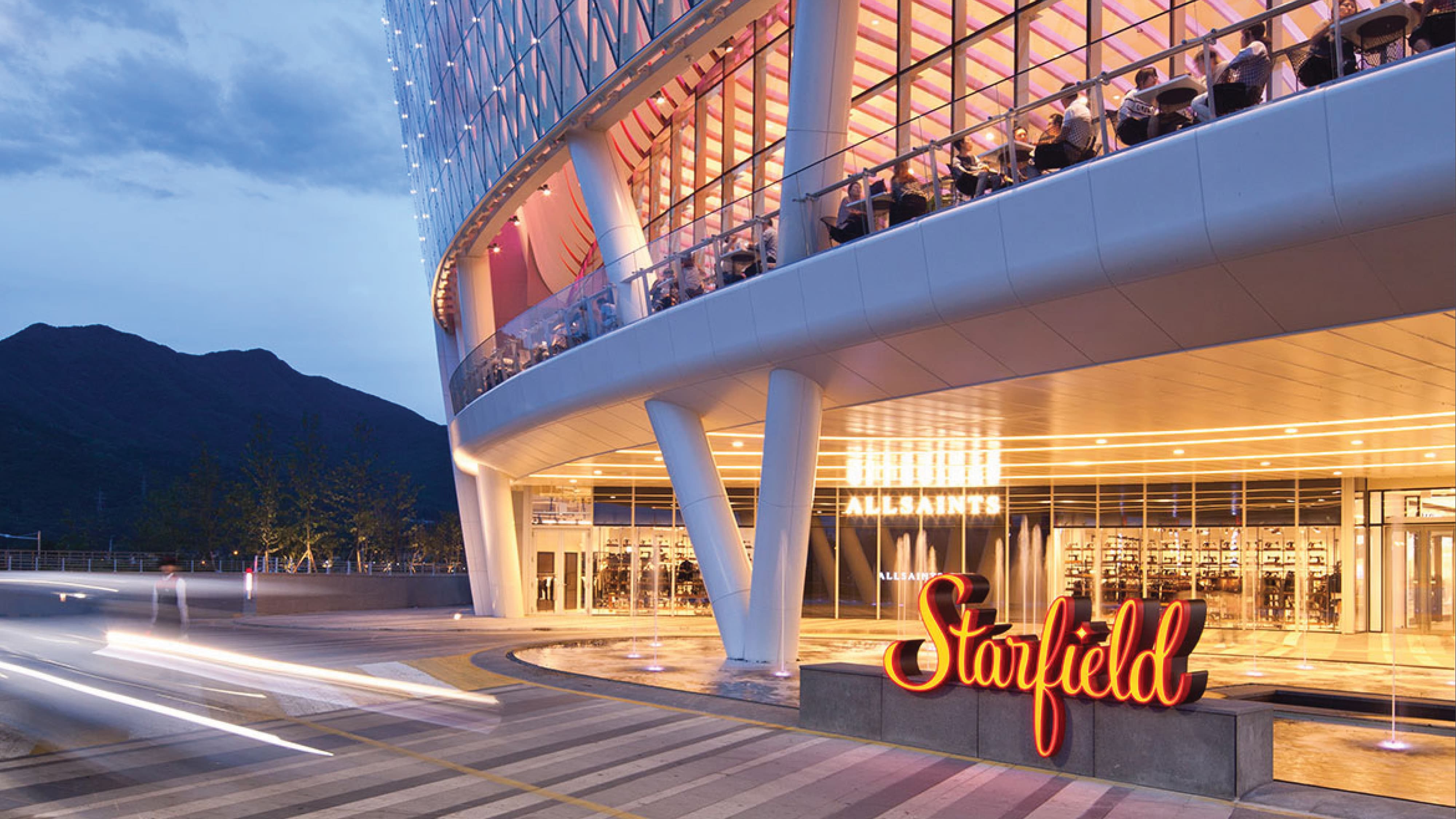 Nighttime exterior of the Starfield mall with an illuminated glass tower displaying the red 'Starfield' script logo, storefronts including Starbucks, and light trails from passing traffic in the foreground.