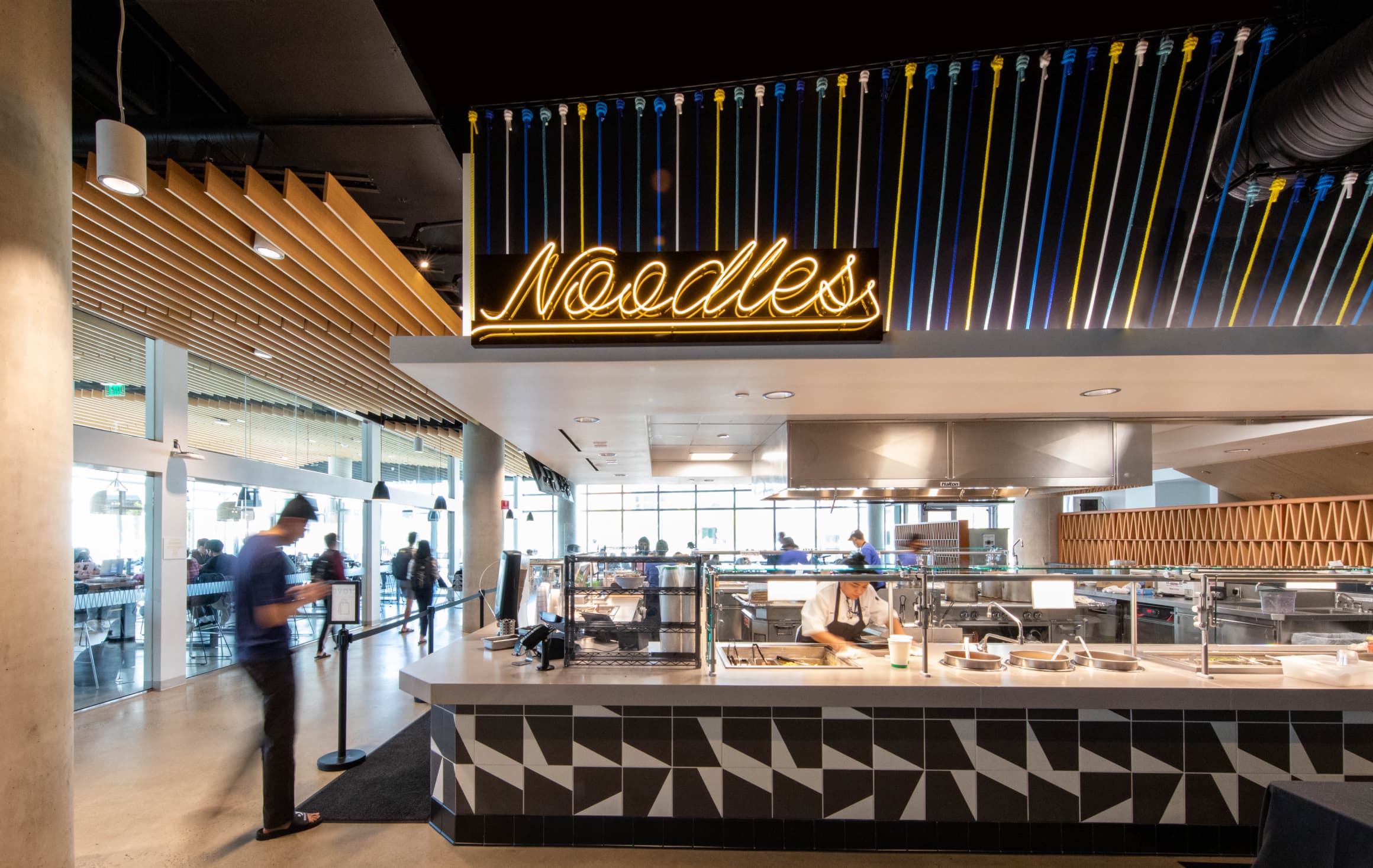 Neon tube lettering sign spelling "Noodles" in a warm light mounted to canopy above vendor in UC San Diego Sixth College's dining hall by RSM Design in San Diego, California. 