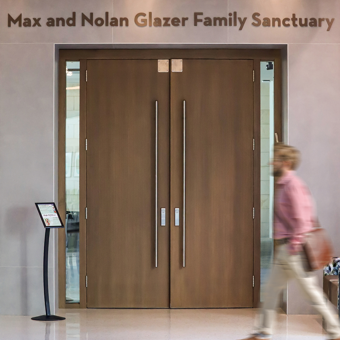 Wood double doors beneath dimensional lettering reading 'Max and Nolan Glazer Family Sanctuary,' with a sign stand beside them and a blurred man walking past in the foreground.