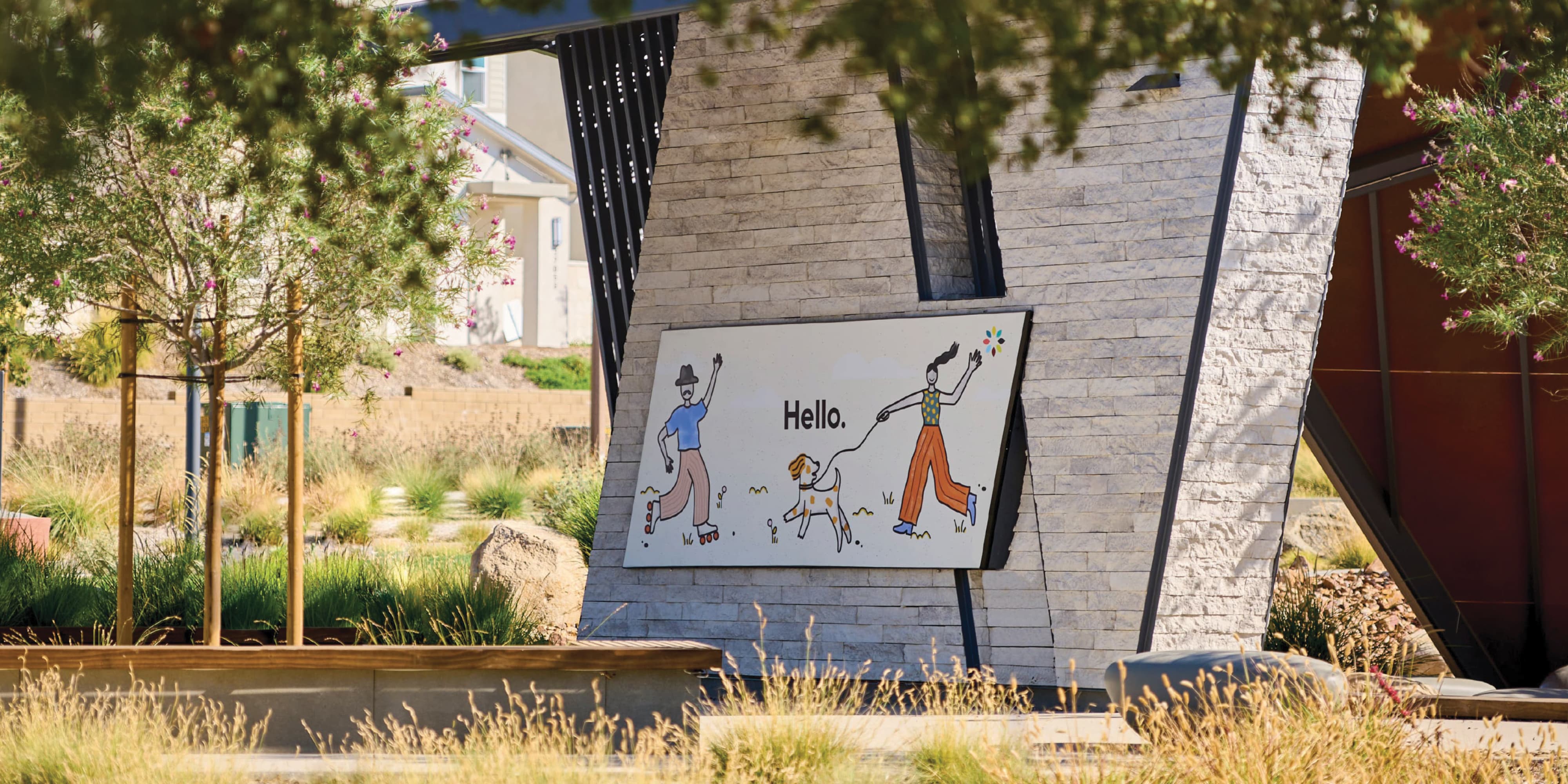 Illustrated 'Hello.' sign depicting a rollerblading man and a woman walking a spotted dog, mounted on an angled stone monument structure, surrounded by native grasses and flowering shrubs with homes visible in the background.