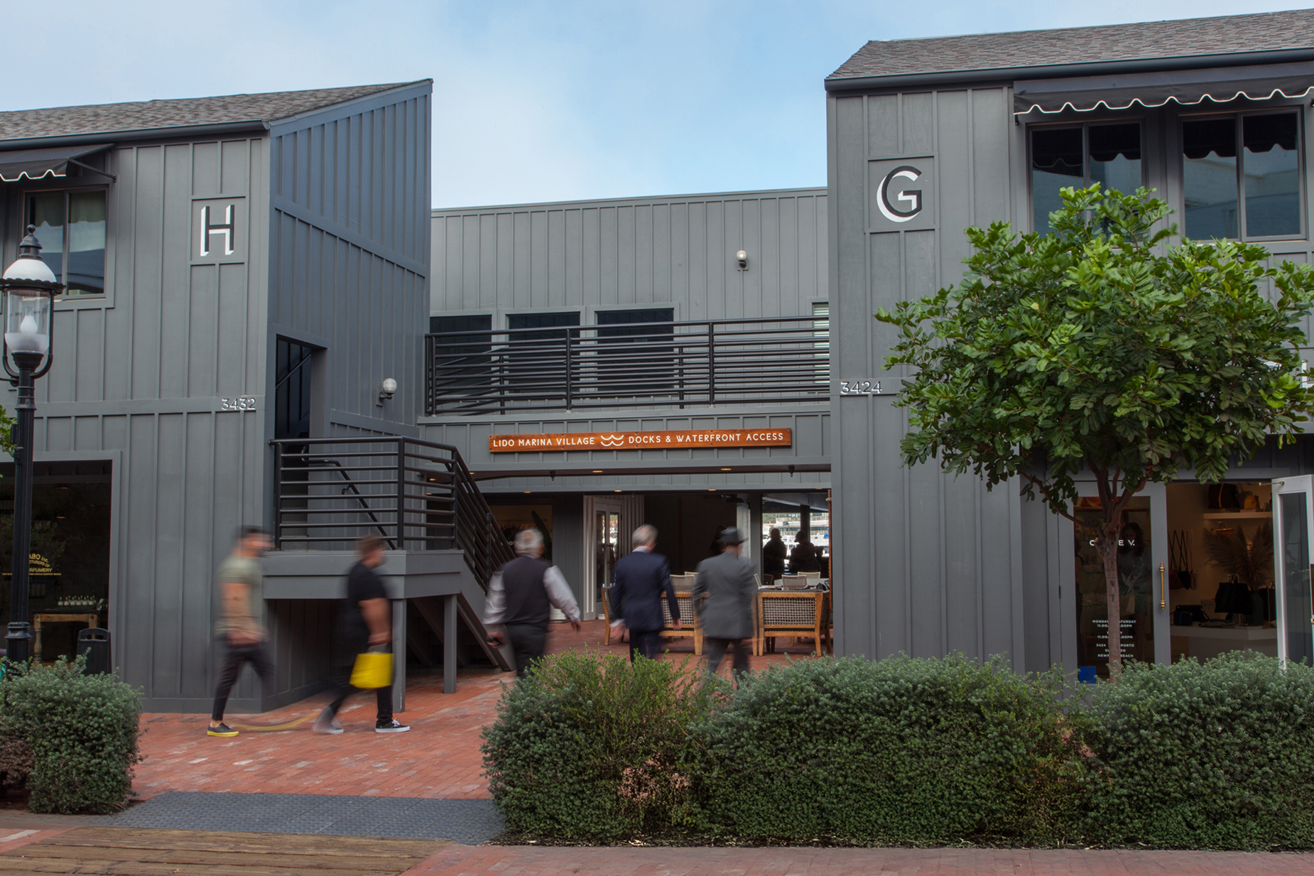 Shoppers walking toward Lido Marina Village Docks and Waterfront Access passage between Buildings G and H with orange wayfinding sign, dark board-and-batten architecture and brick pathway in Newport Beach, California